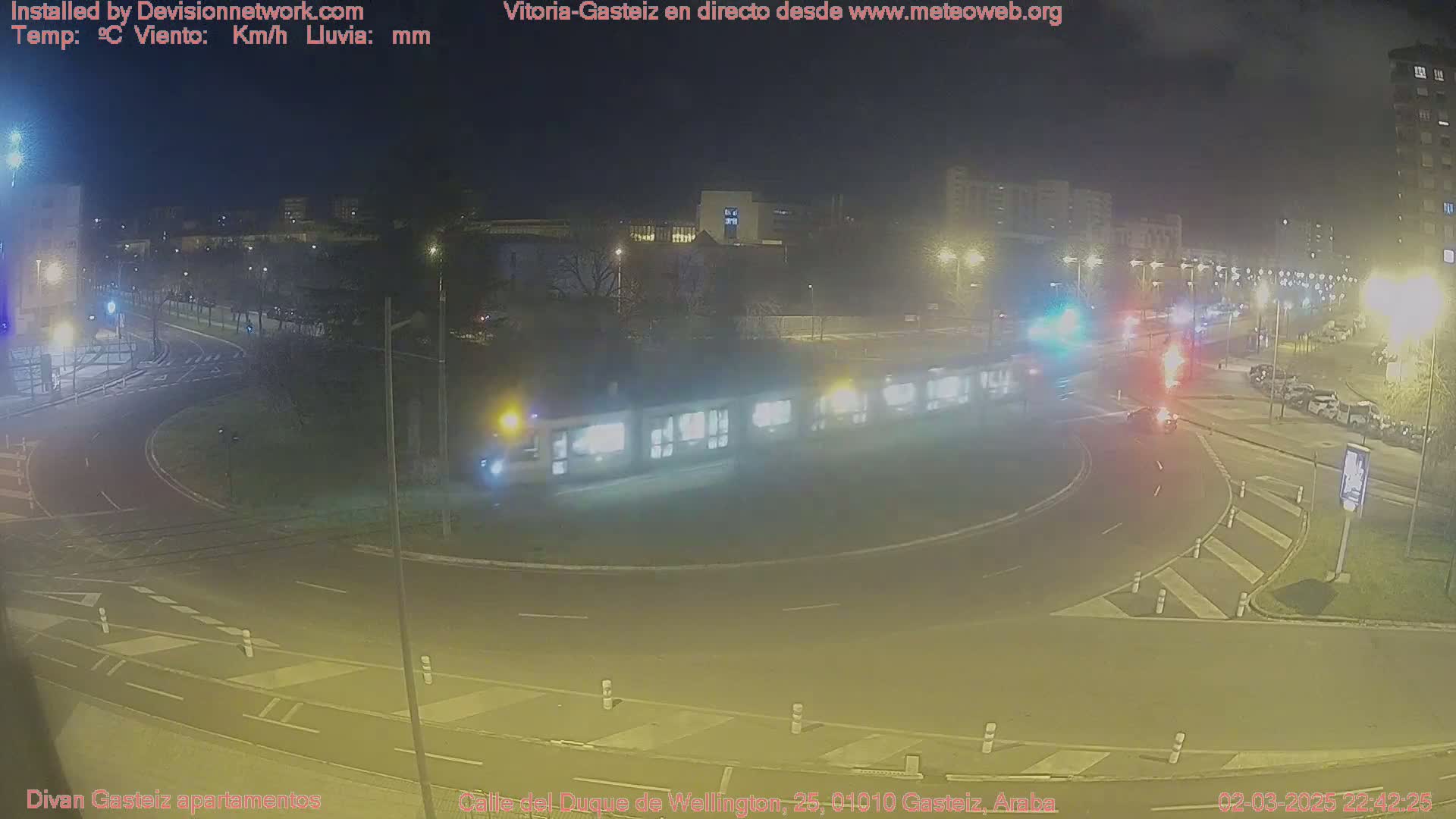 At night, a long tram travels through a roundabout intersection with cars and streetlights illuminating the scene.