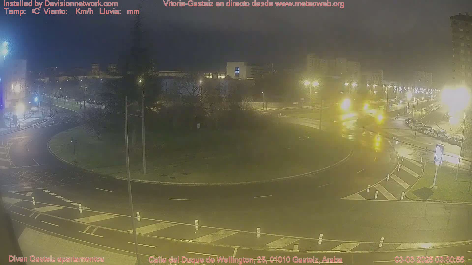 A wet, nighttime view of a roundabout and surrounding streets with sparse traffic and buildings in the background under streetlights.