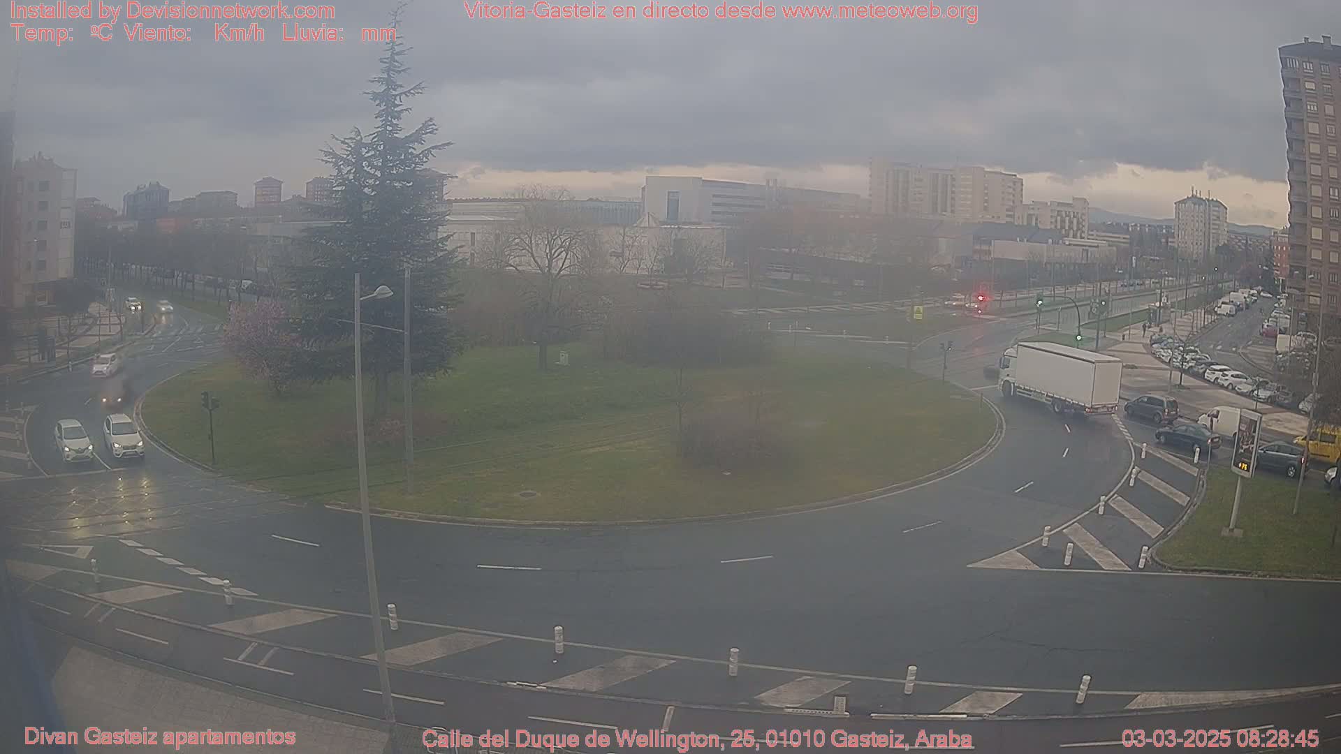 A mostly overcast day shows a traffic circle with several cars and a truck, surrounded by buildings and trees.