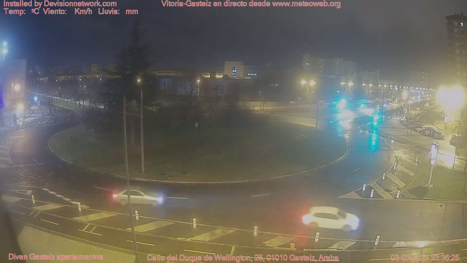 A wet, nighttime scene shows a roundabout with several cars driving through it, under streetlights in a city.