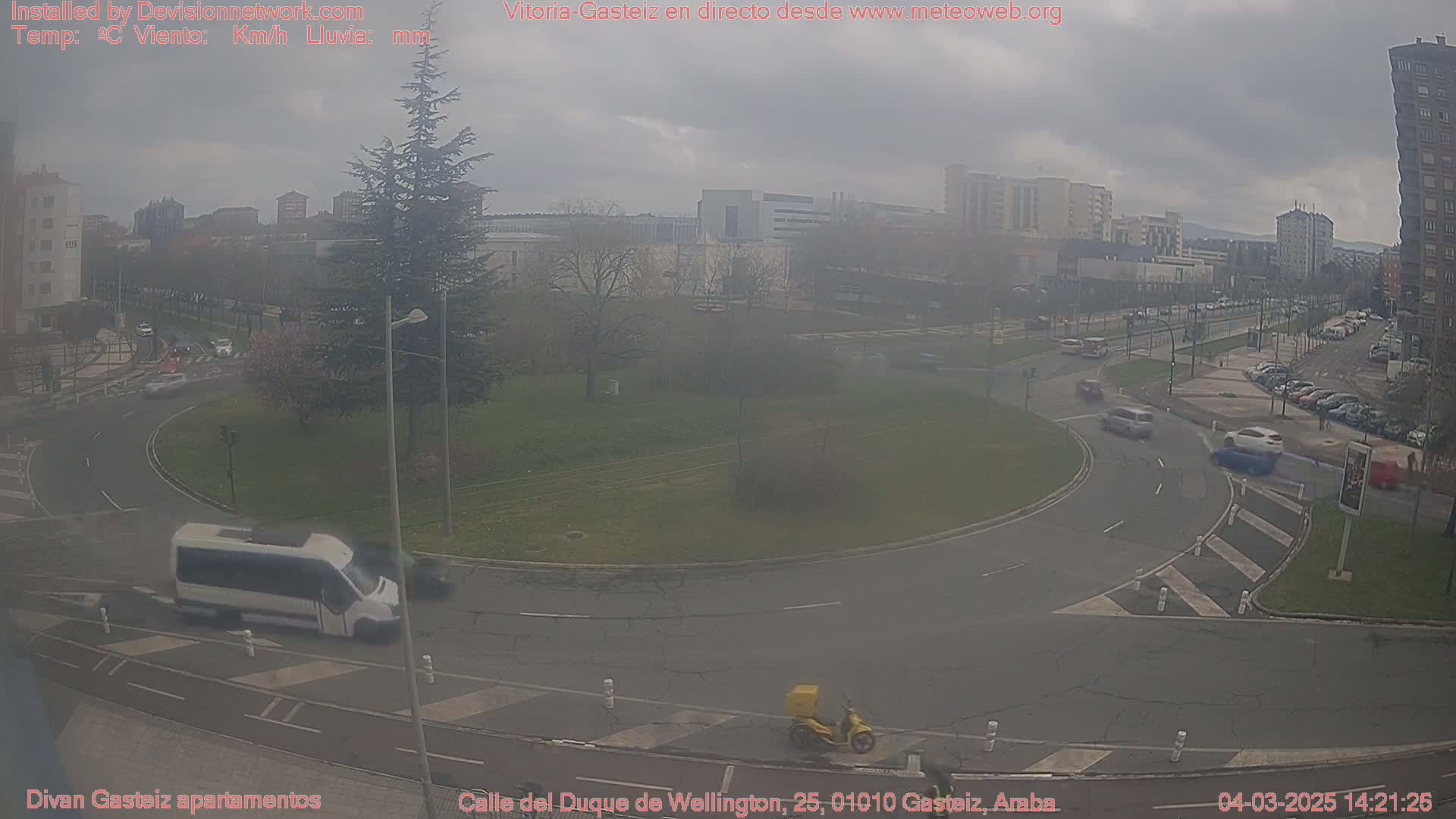 A hazy overcast day reveals a roundabout in a city with several cars and a van driving around it, and a delivery person on a scooter.