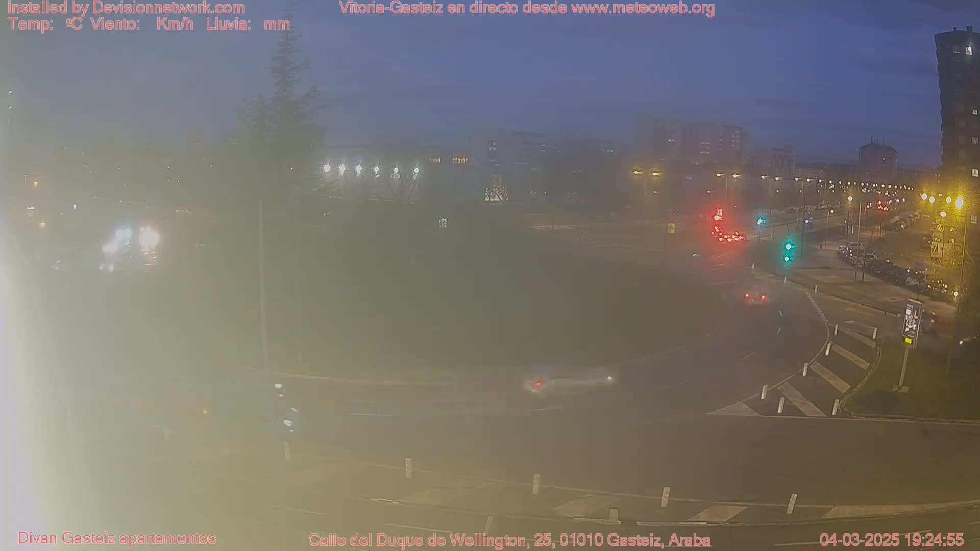 A hazy, night-time view of a traffic circle with cars and streetlights, under a dark, twilight sky.