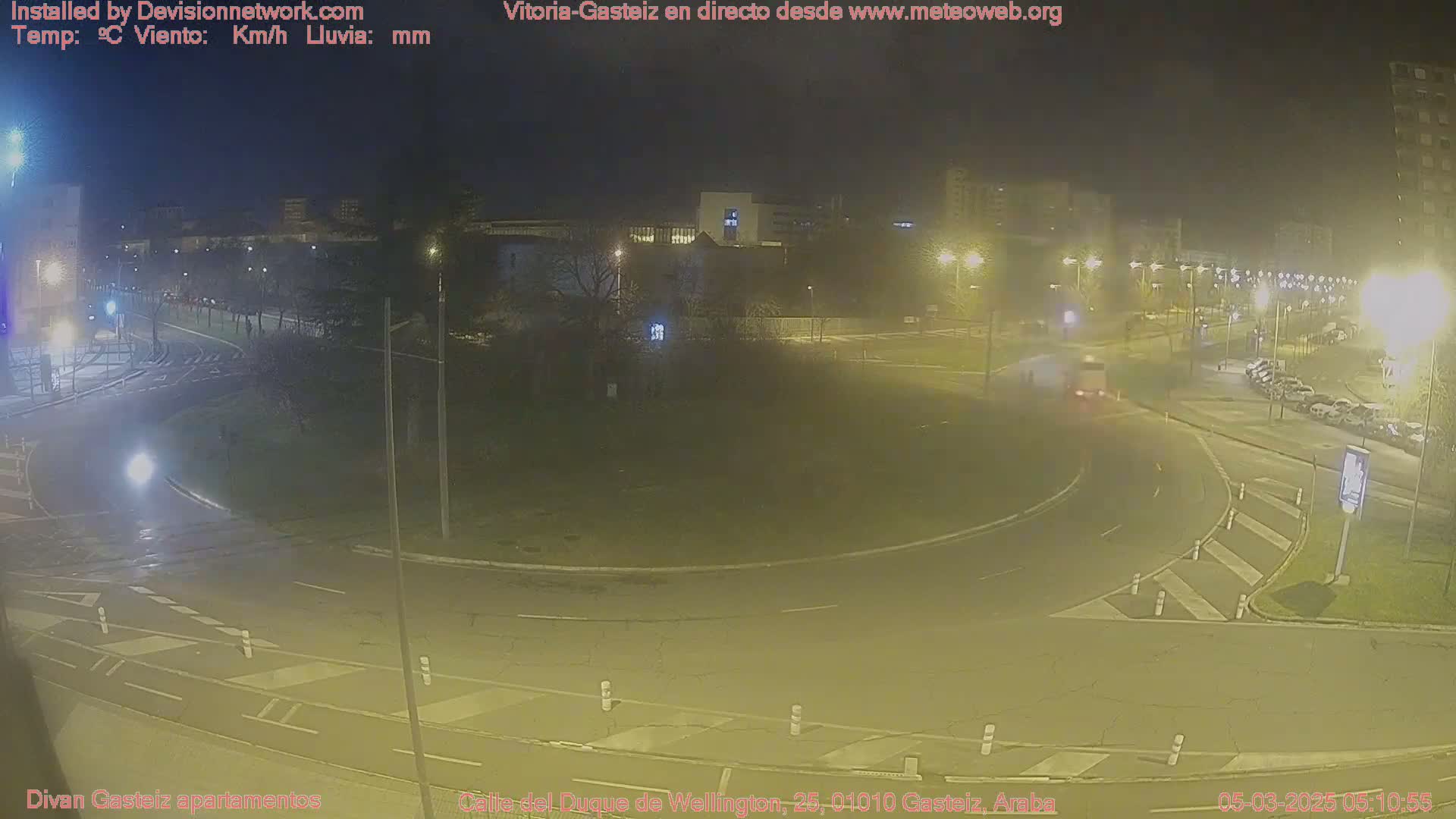 A nighttime view of a roundabout with a few cars and streetlights illuminating a mostly empty roadway.