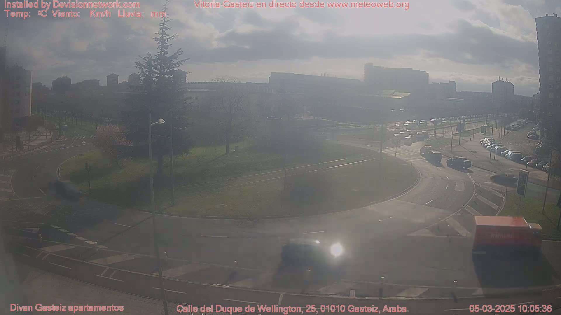 A hazy, sunny day reveals a roundabout and surrounding city streets with several vehicles visible, buildings in the background.