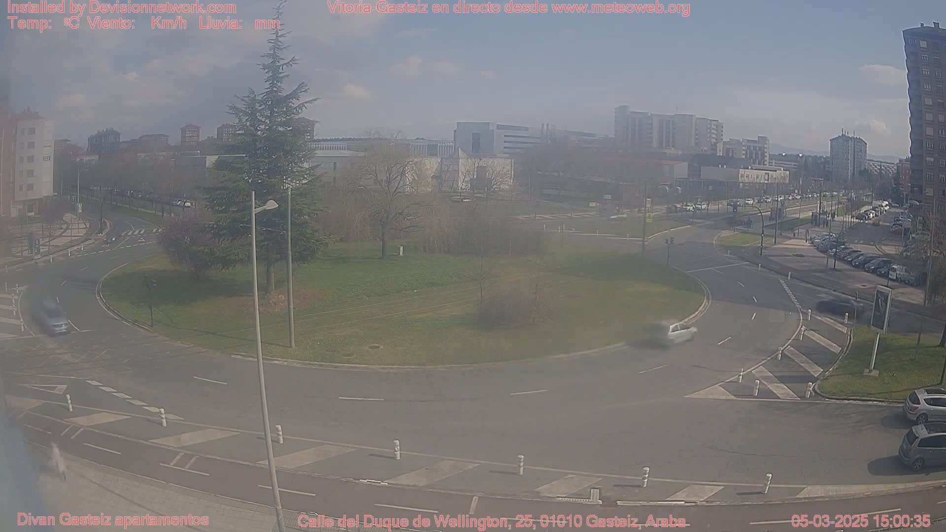 A hazy, sunny day reveals a roundabout intersection in a city with several cars and buildings in the background.