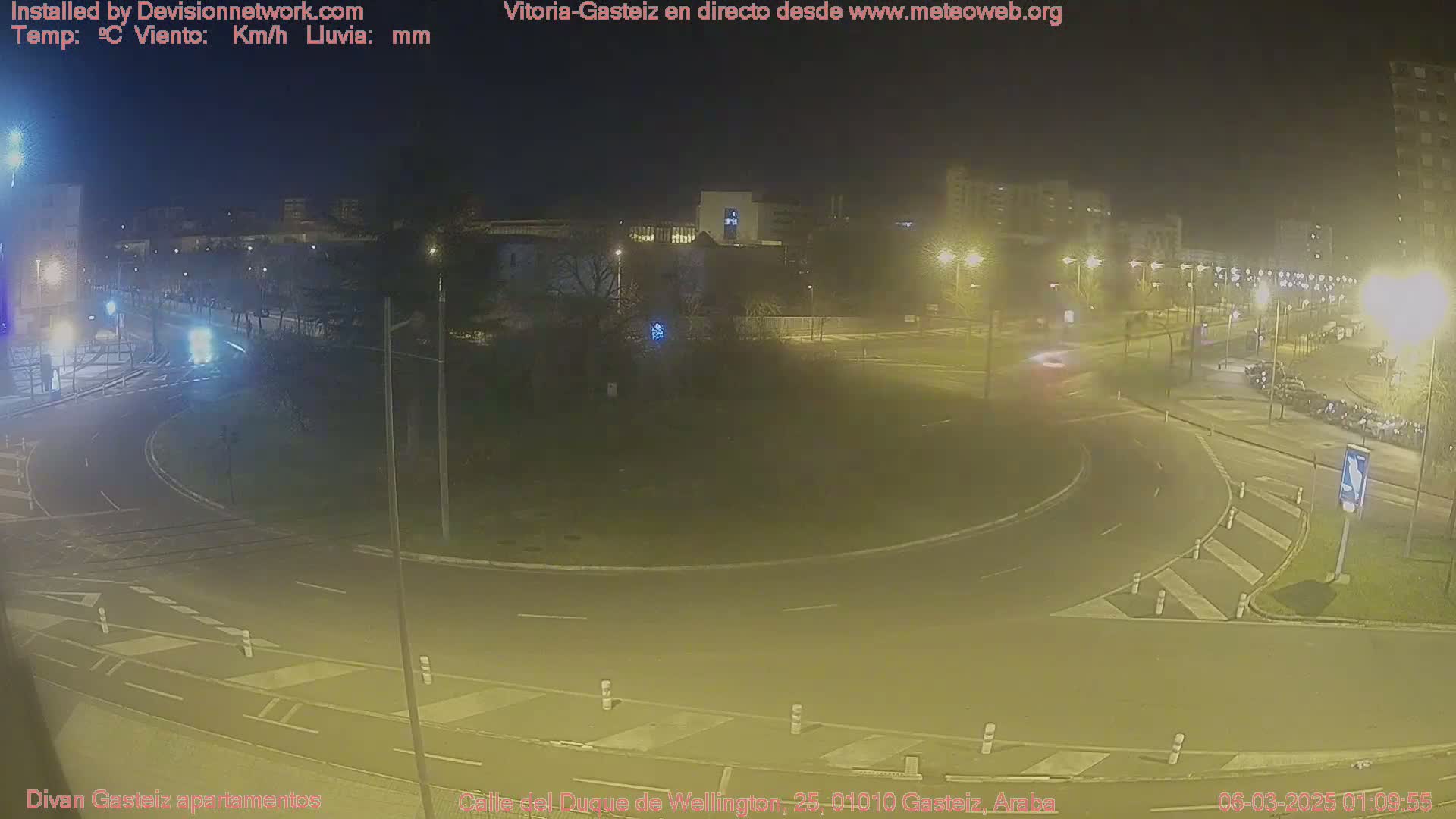 A nighttime view of a traffic roundabout in a city, with several vehicles visible and streetlights illuminating the scene.