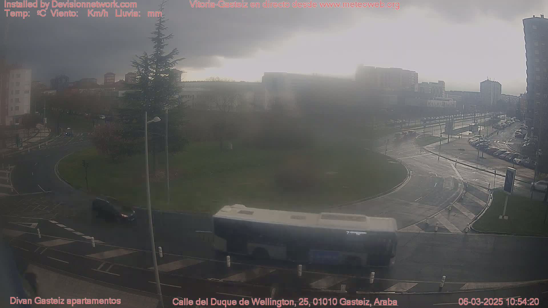 A hazy, overcast day reveals a roundabout and surrounding streets with several cars and a bus, near multi-story buildings.