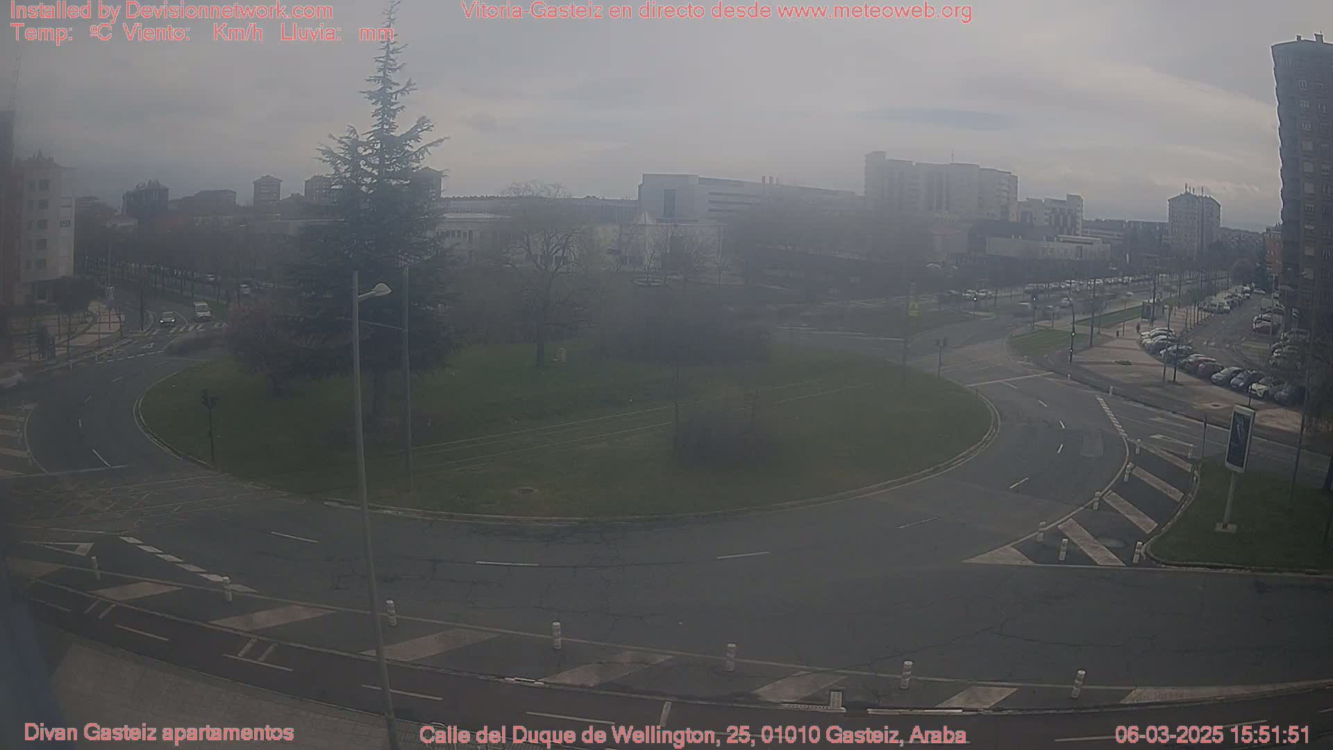 A hazy, overcast day reveals a roundabout surrounded by a city street scene with buildings and parked cars.