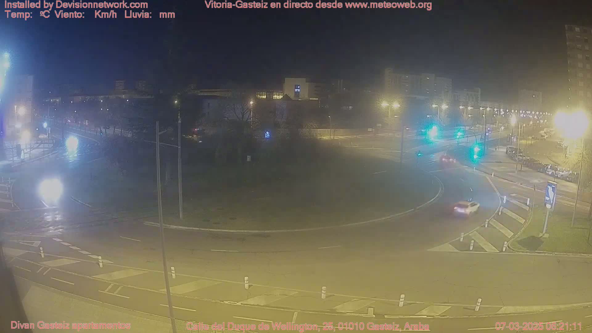 A wet, nighttime scene shows a roundabout with several cars and buildings in the background under streetlights.