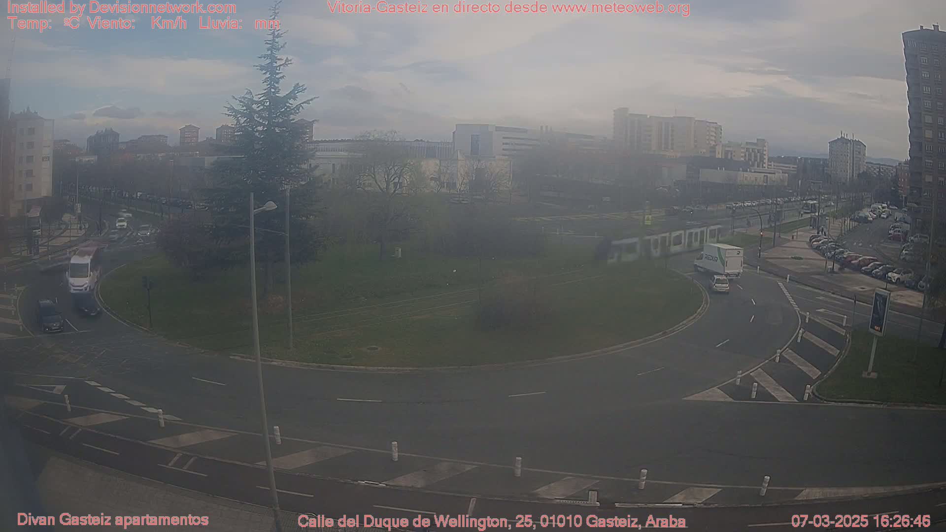 A hazy, overcast day shows a roundabout with several cars and trucks navigating it, surrounded by buildings and a grassy area with trees.