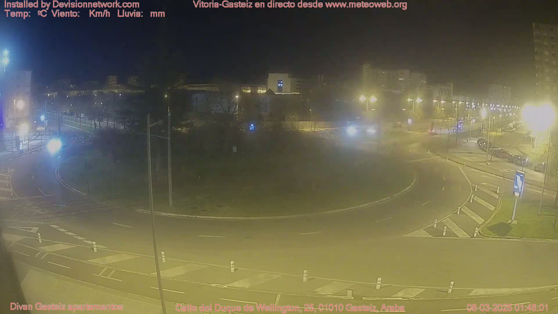 A nighttime view of a roundabout with cars and buildings in the background under artificial lighting.