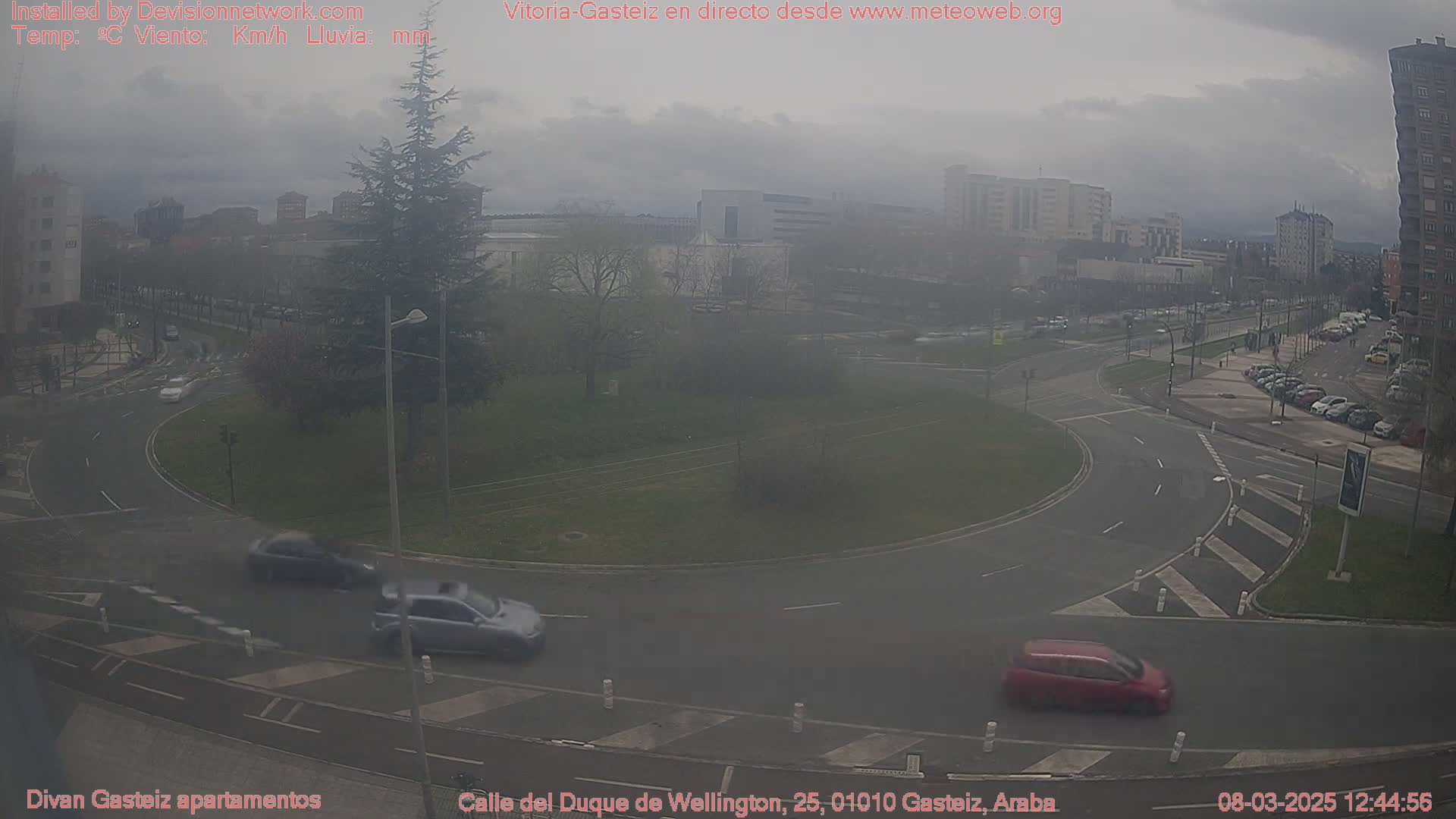 A hazy, overcast day reveals a roundabout with several cars, surrounded by buildings and a small grassy area.
