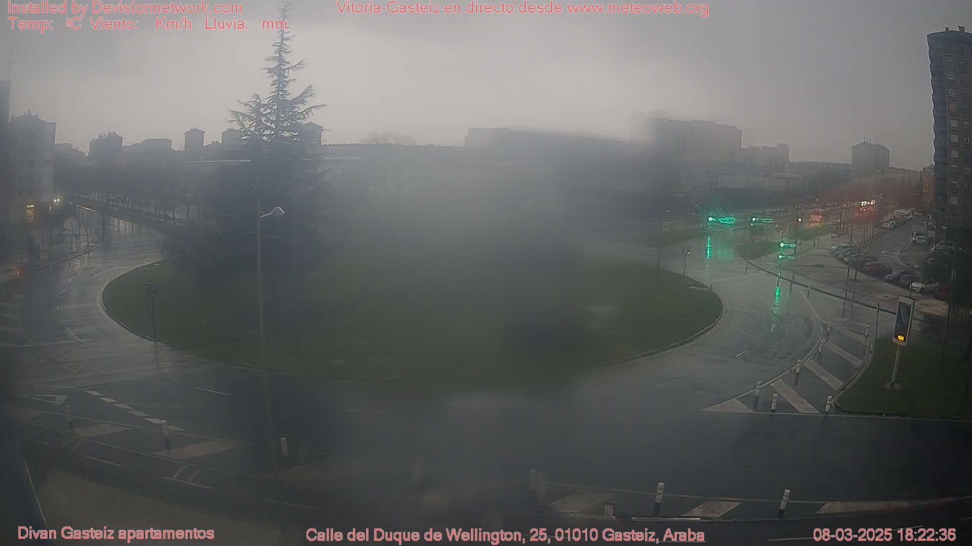 A foggy, rainy scene shows a wet roundabout and surrounding streets in a city, with buildings visible in the background.