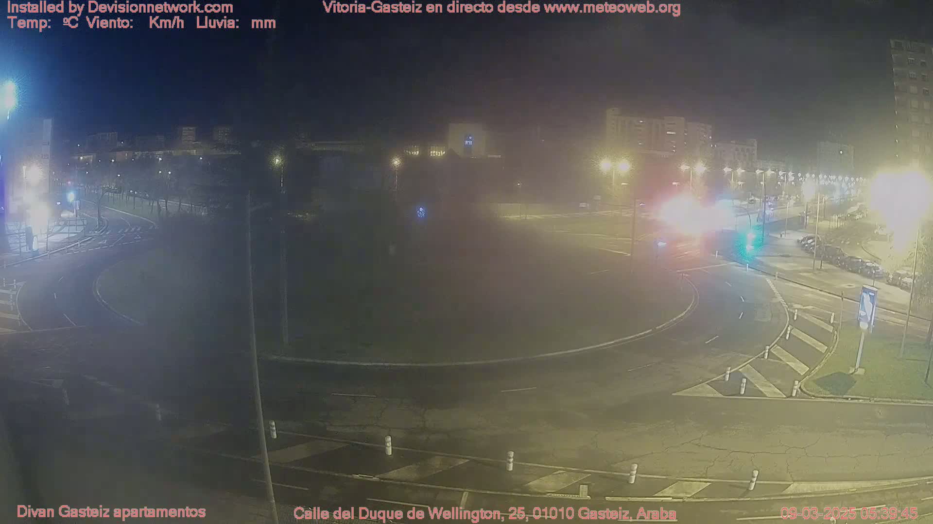 A nighttime, hazy view of a roundabout and surrounding streets with some cars and buildings visible under streetlights.