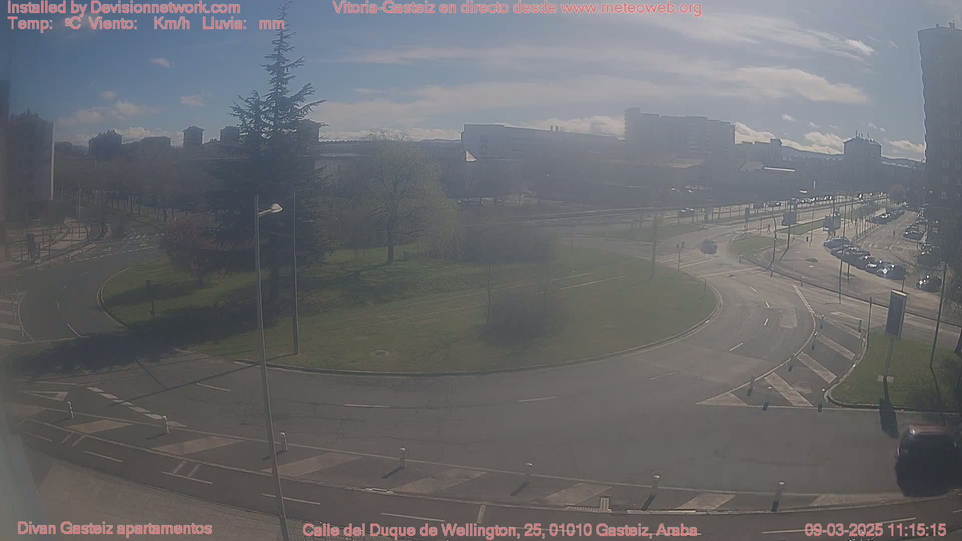 A hazy sunny day reveals a roundabout and roadways with parked cars and light traffic, surrounded by buildings and green spaces.