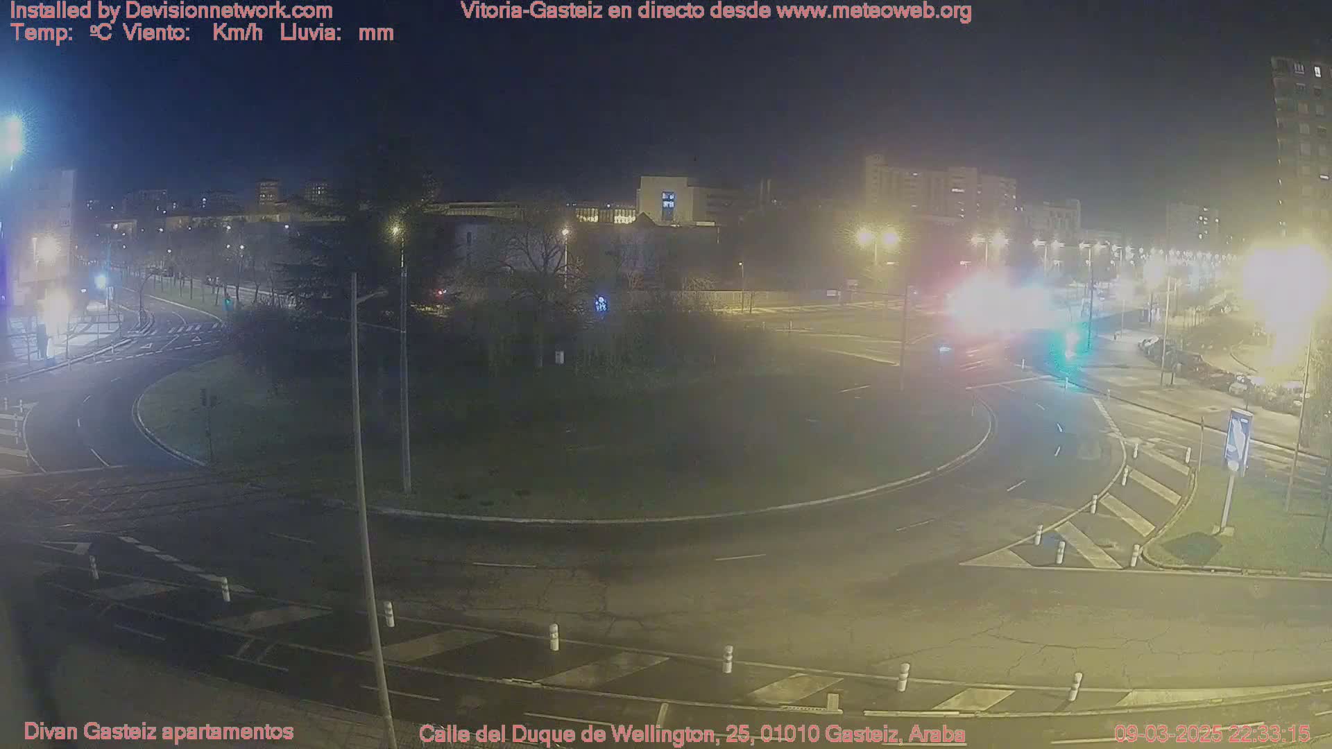 A nighttime view of a roundabout intersection with several buildings and streetlights visible in the background.