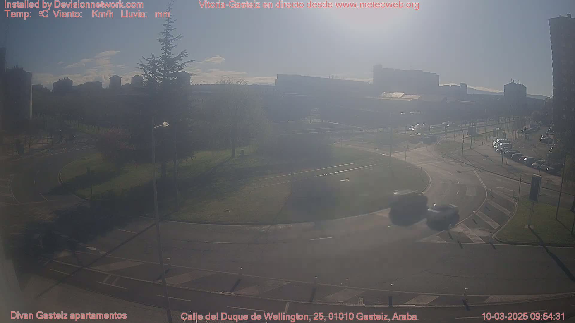 A hazy, sunny day reveals a roundabout and surrounding roadways with sparse traffic, buildings in the distance, and a small park with a tree in the foreground.