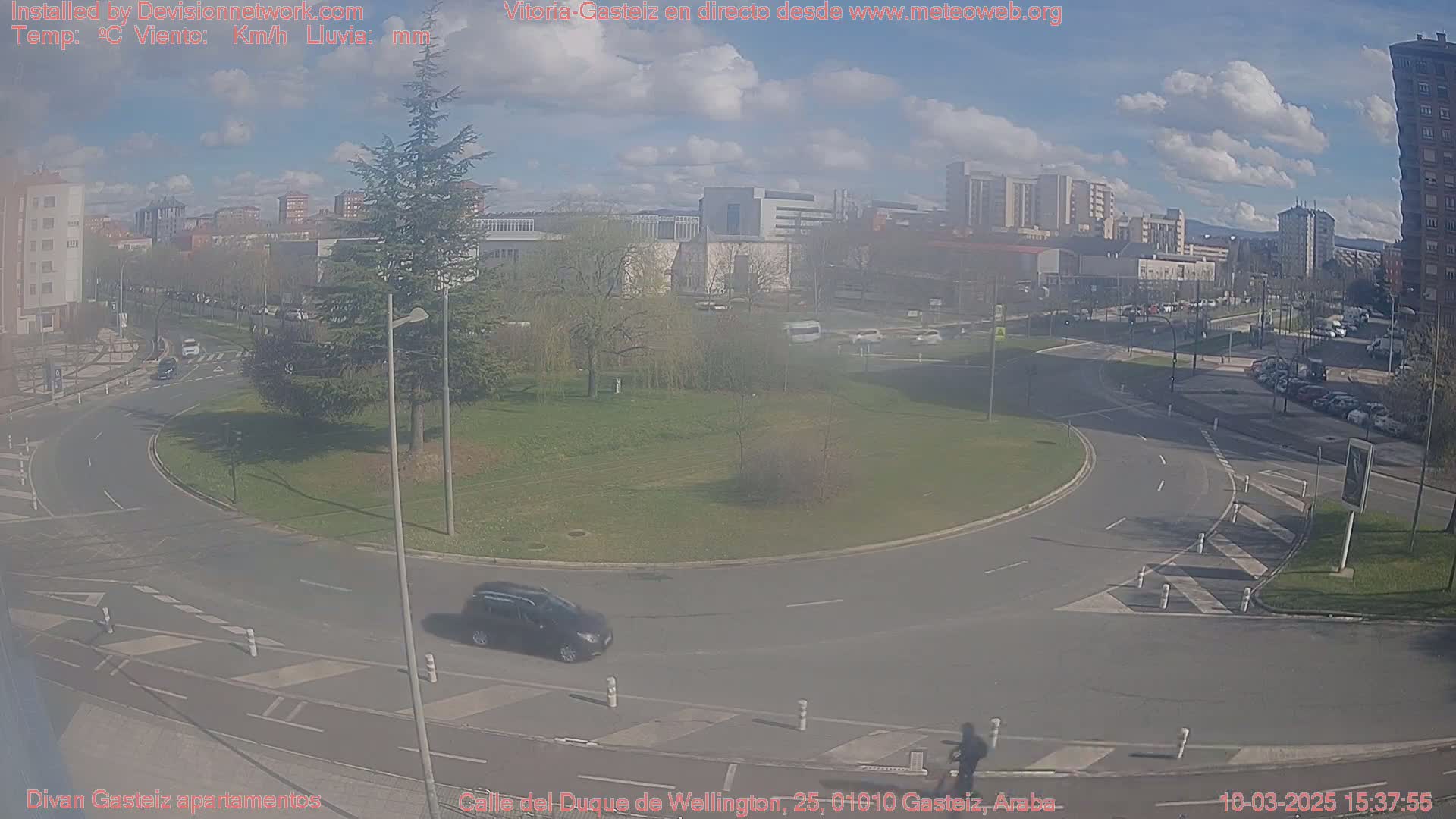 A partly cloudy day shows a roundabout with a few cars and a pedestrian, surrounded by buildings and a grassy area.