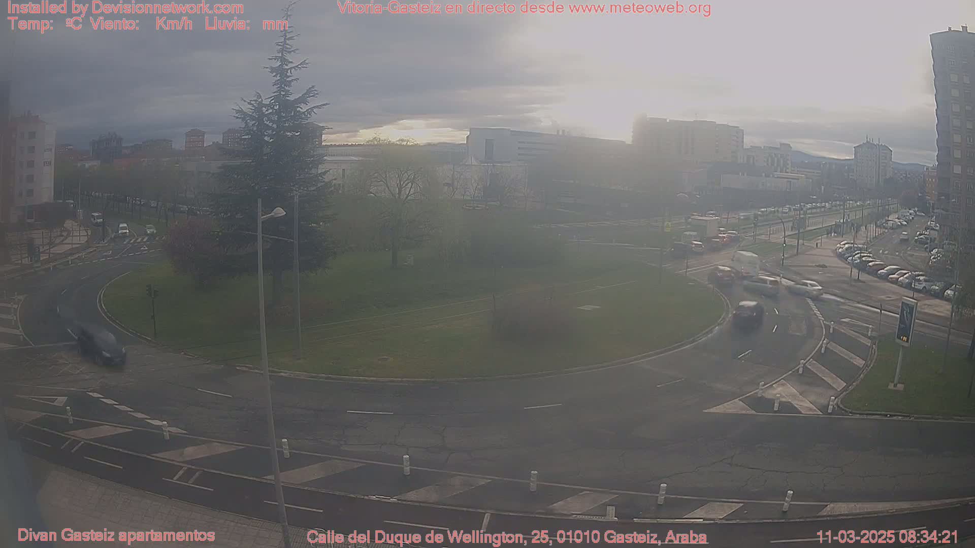 A hazy, overcast day reveals a roundabout with several cars and a small grassy area, surrounded by buildings and parked cars.