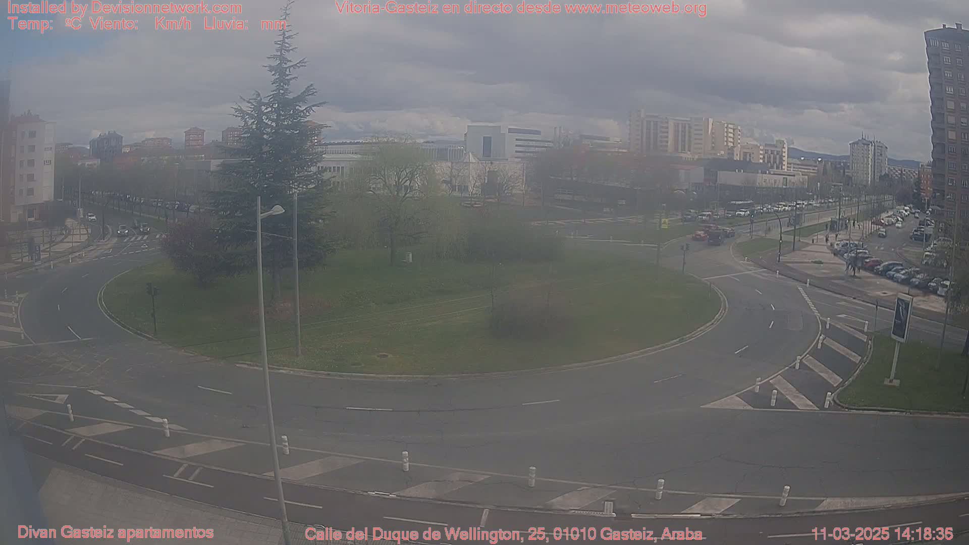 A hazy view of a roundabout and surrounding city streets, with several buildings and parked cars visible in the background.