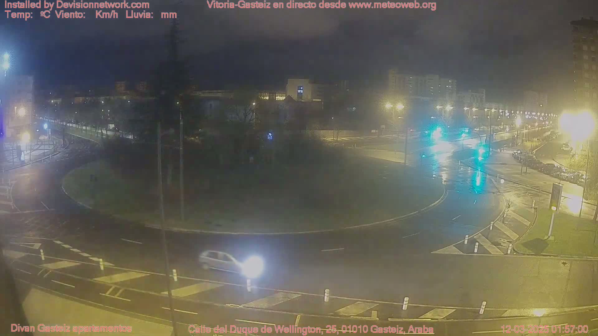 A nighttime, wet roundabout with several cars and surrounding buildings under streetlights.