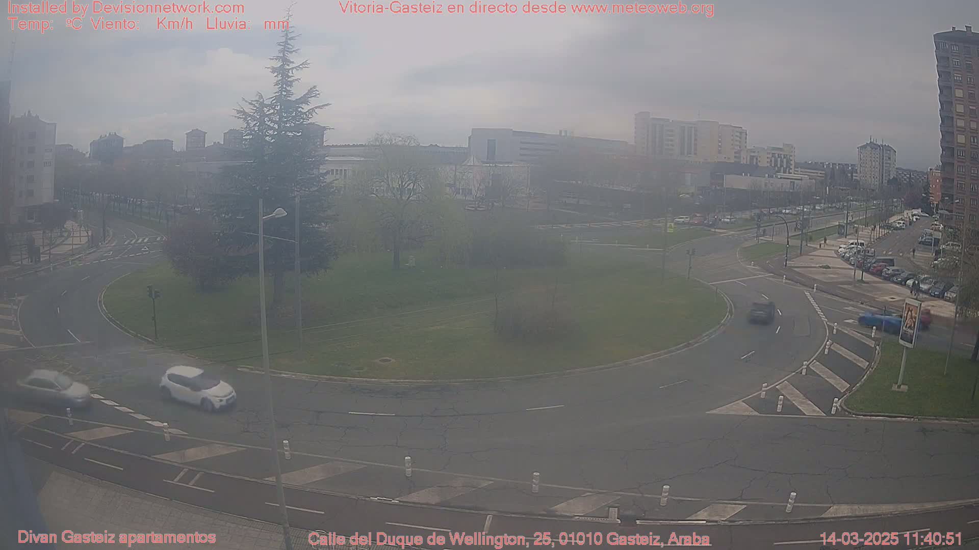 A hazy, overcast day reveals a roundabout in a city, with several cars visible and buildings in the background.