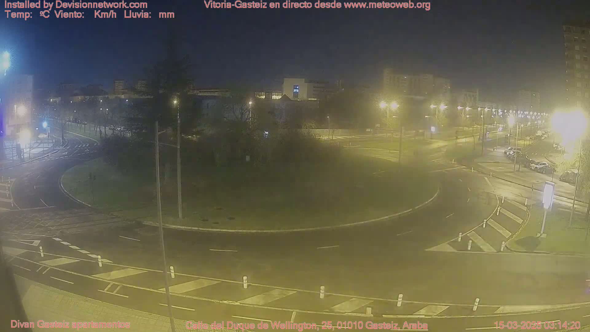 A nighttime view of a wet roundabout and surrounding roads with parked cars and buildings in the distance under streetlights.