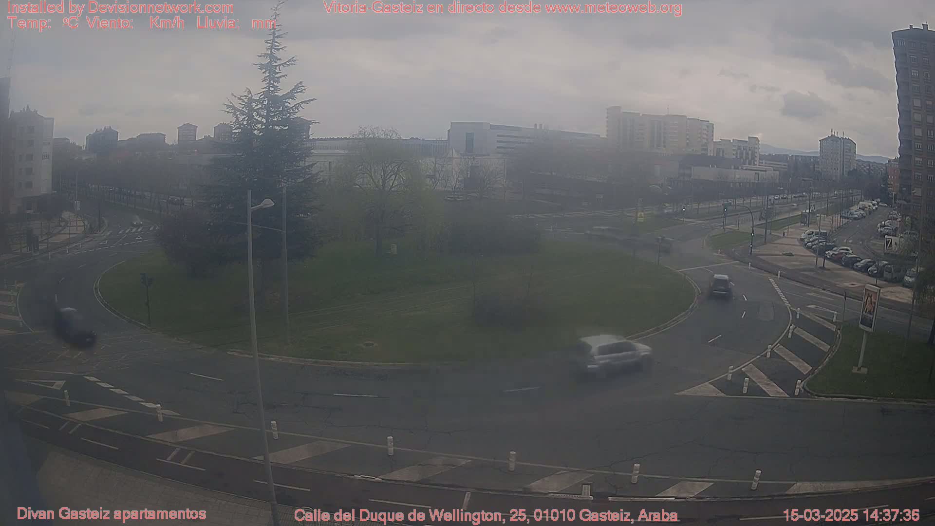 A hazy overcast day reveals a roundabout in a city with several cars visible, surrounded by buildings and trees.