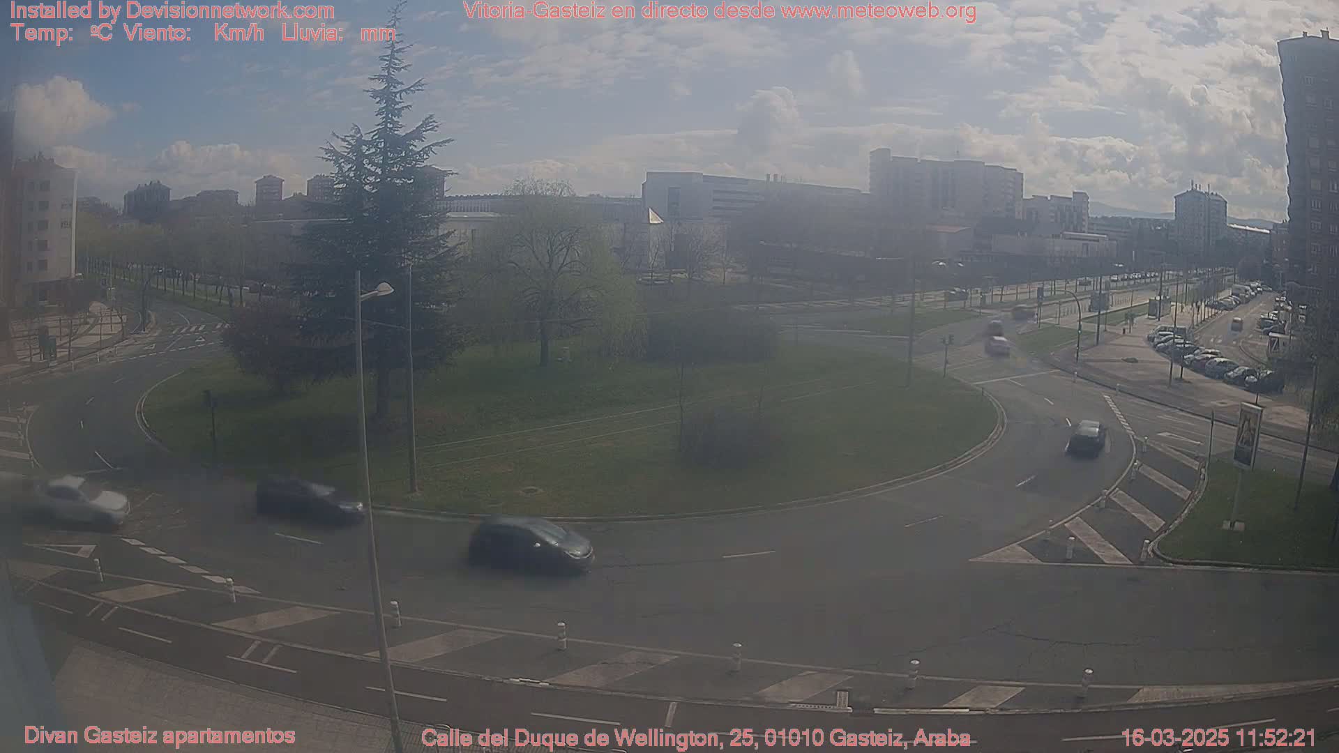 A hazy, partly sunny day reveals a roundabout with several cars and a city skyline in the background.