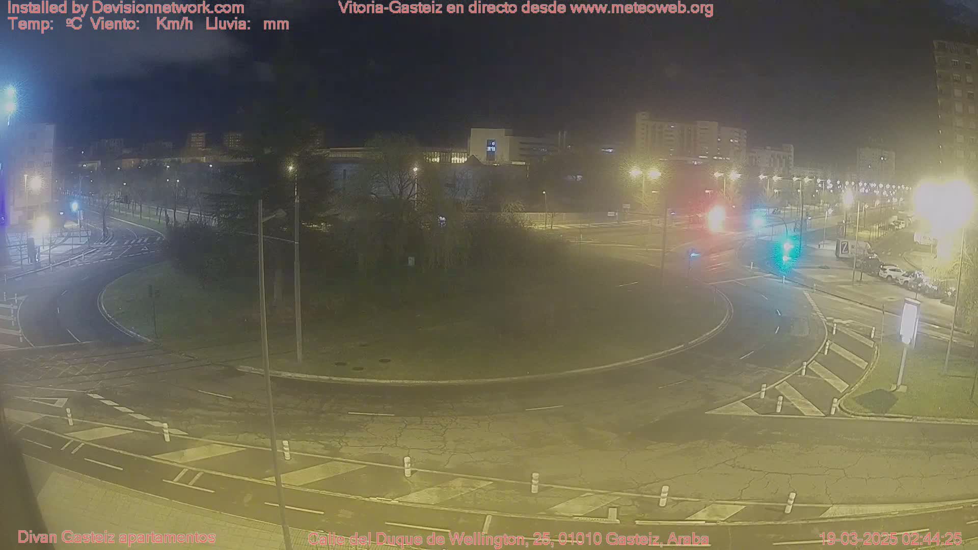 A nighttime view of a roundabout intersection with sparsely trafficked roads and surrounding buildings under a clear, dark sky.