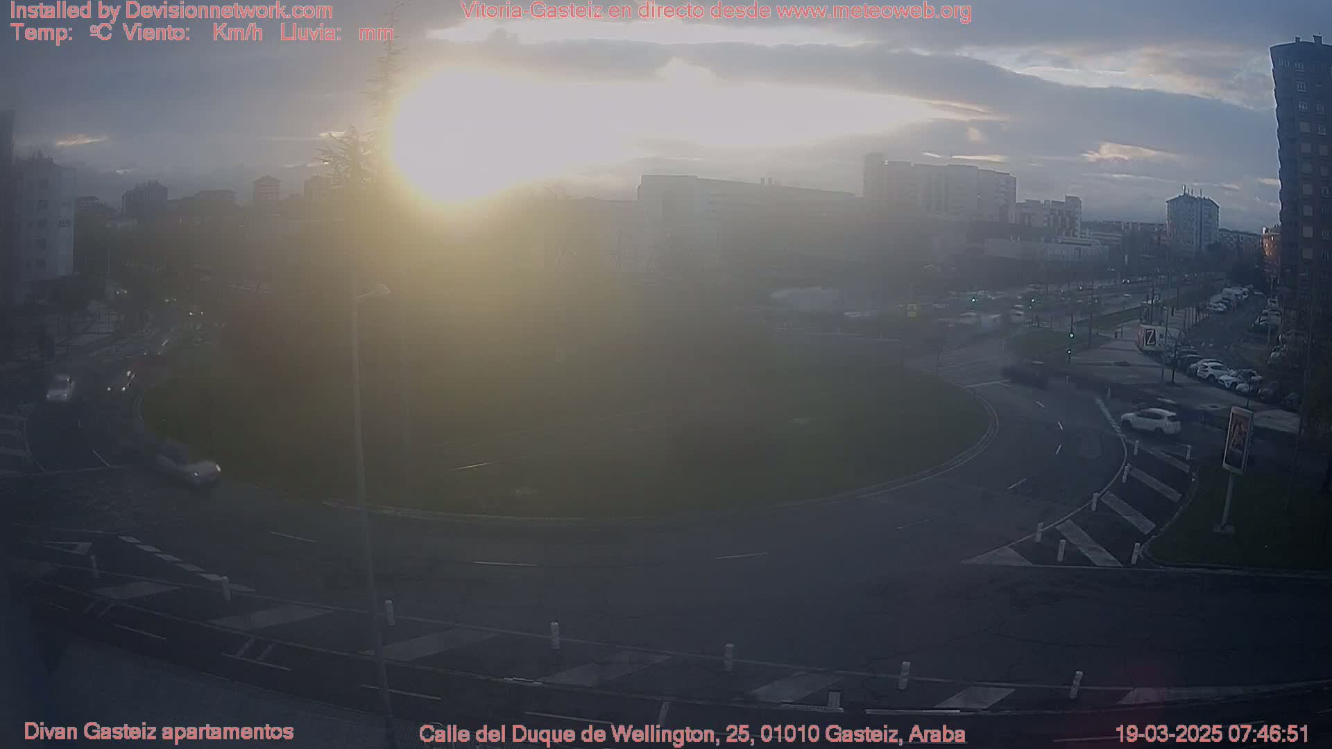 A hazy sunrise illuminates a roundabout with several cars visible, surrounded by buildings in a city setting.