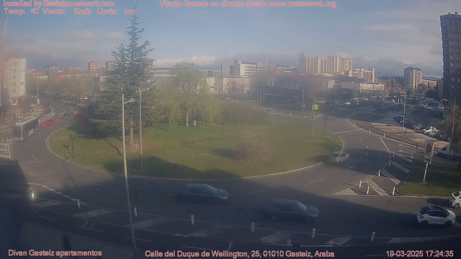 A mostly sunny view of a roundabout in a city with several cars driving around it, some buildings in the background, and a few trees in a nearby park.