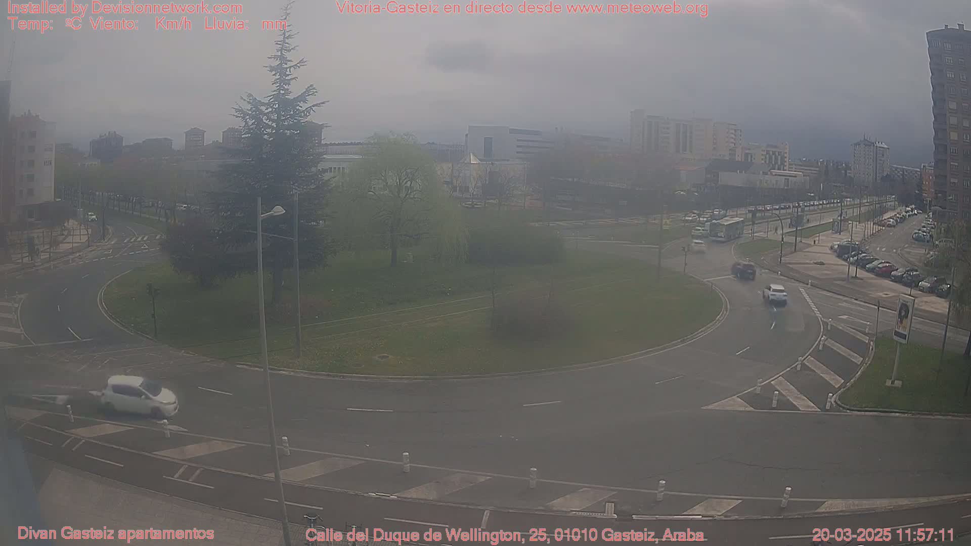 A hazy view of a roundabout in a city, with several cars driving on the roads and buildings visible in the background.