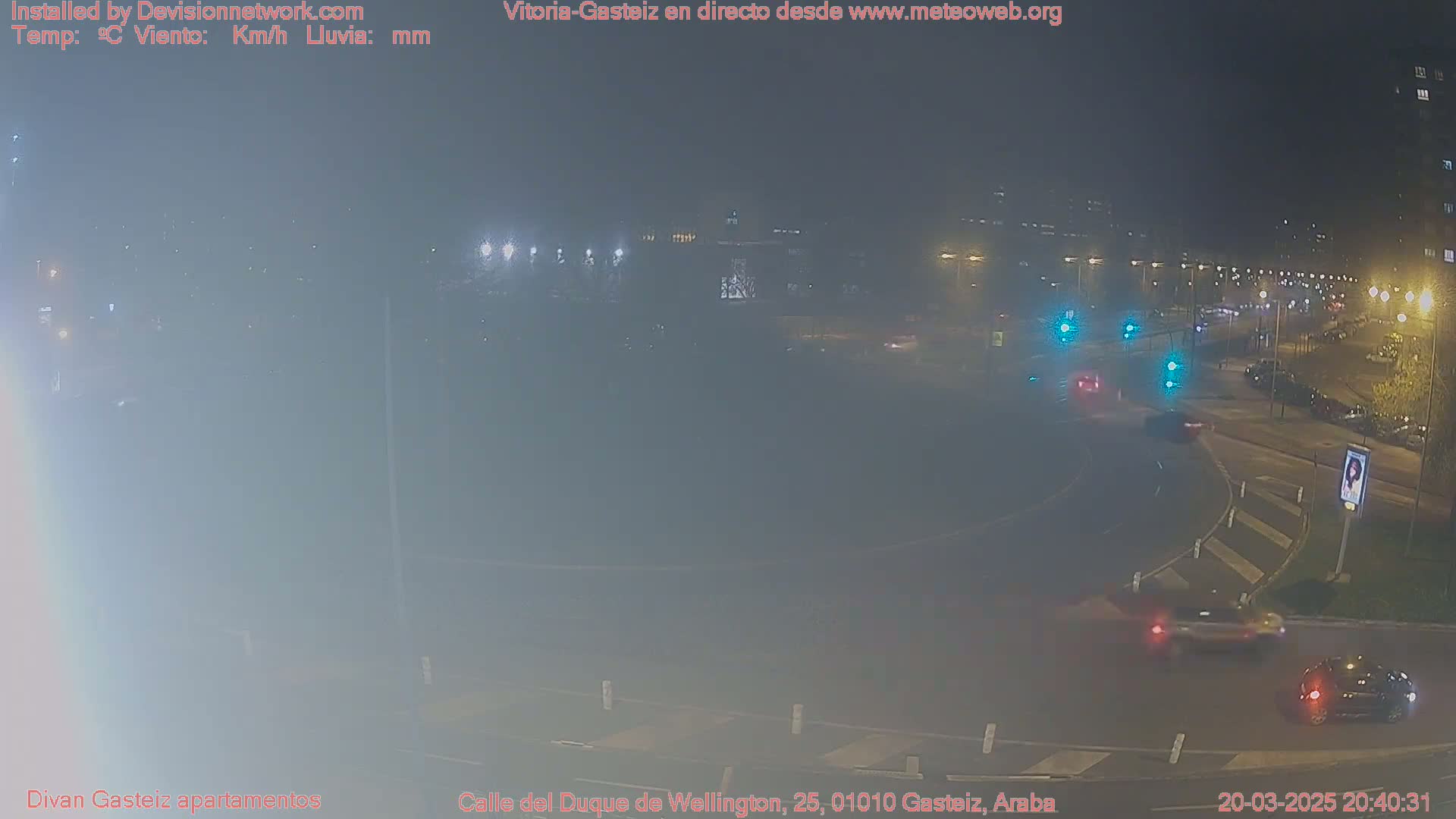 A nighttime, hazy view of a roundabout with several cars visible, and streetlights illuminating the scene.