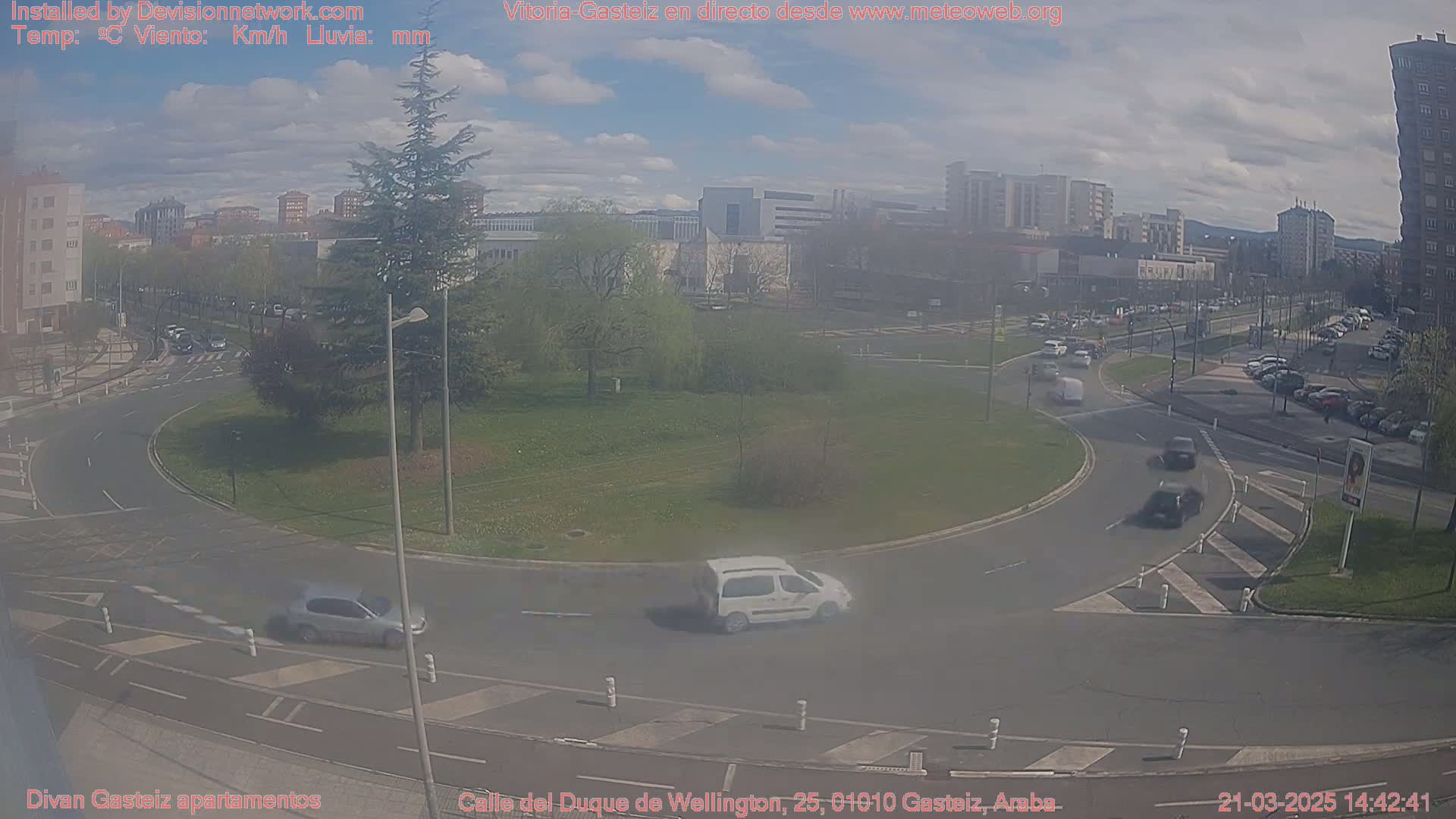 A partly cloudy day reveals a roundabout with several cars driving around it, surrounded by buildings and greenery.