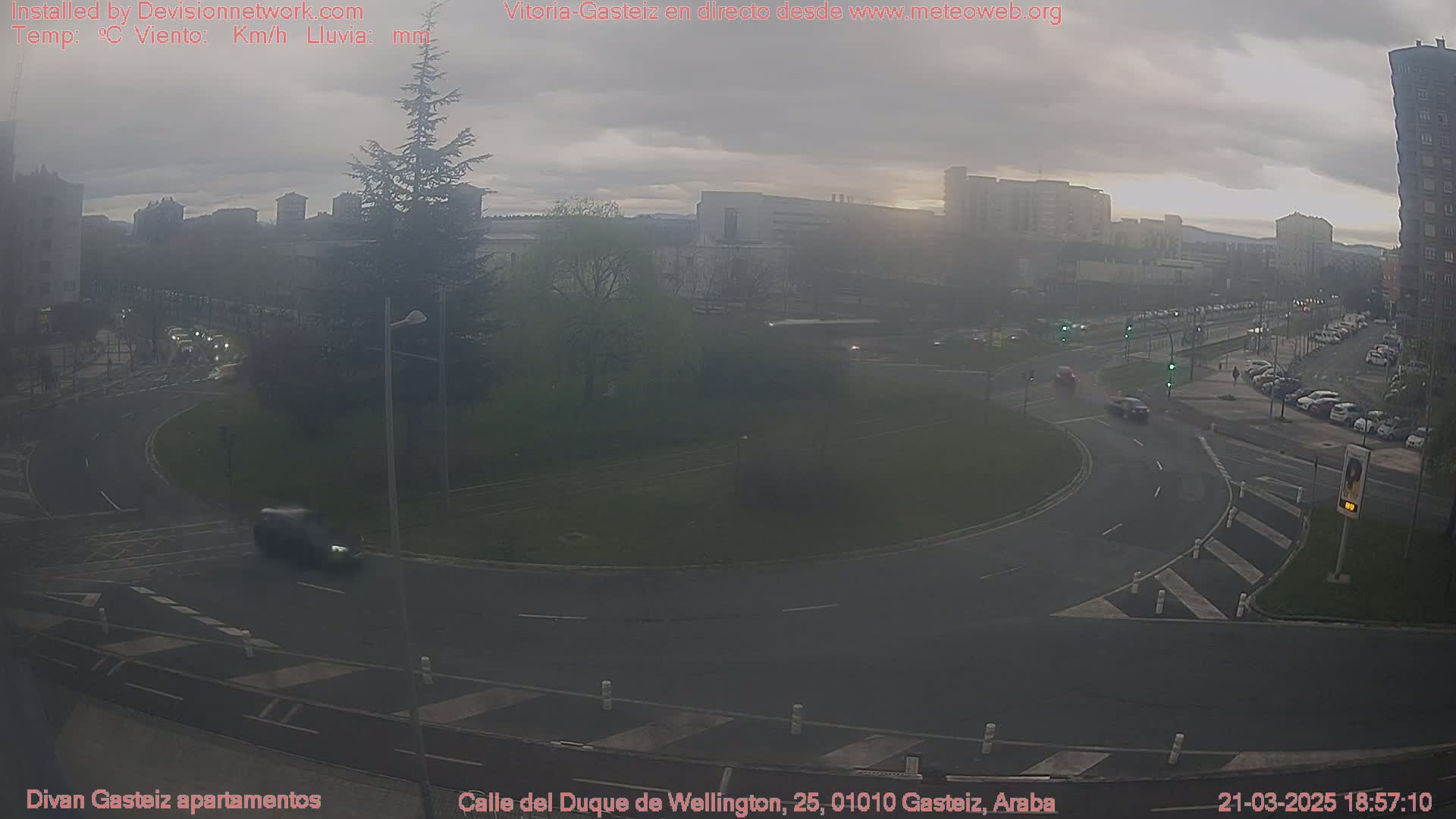 A hazy, overcast scene shows a roundabout with several cars and buildings in the background.
