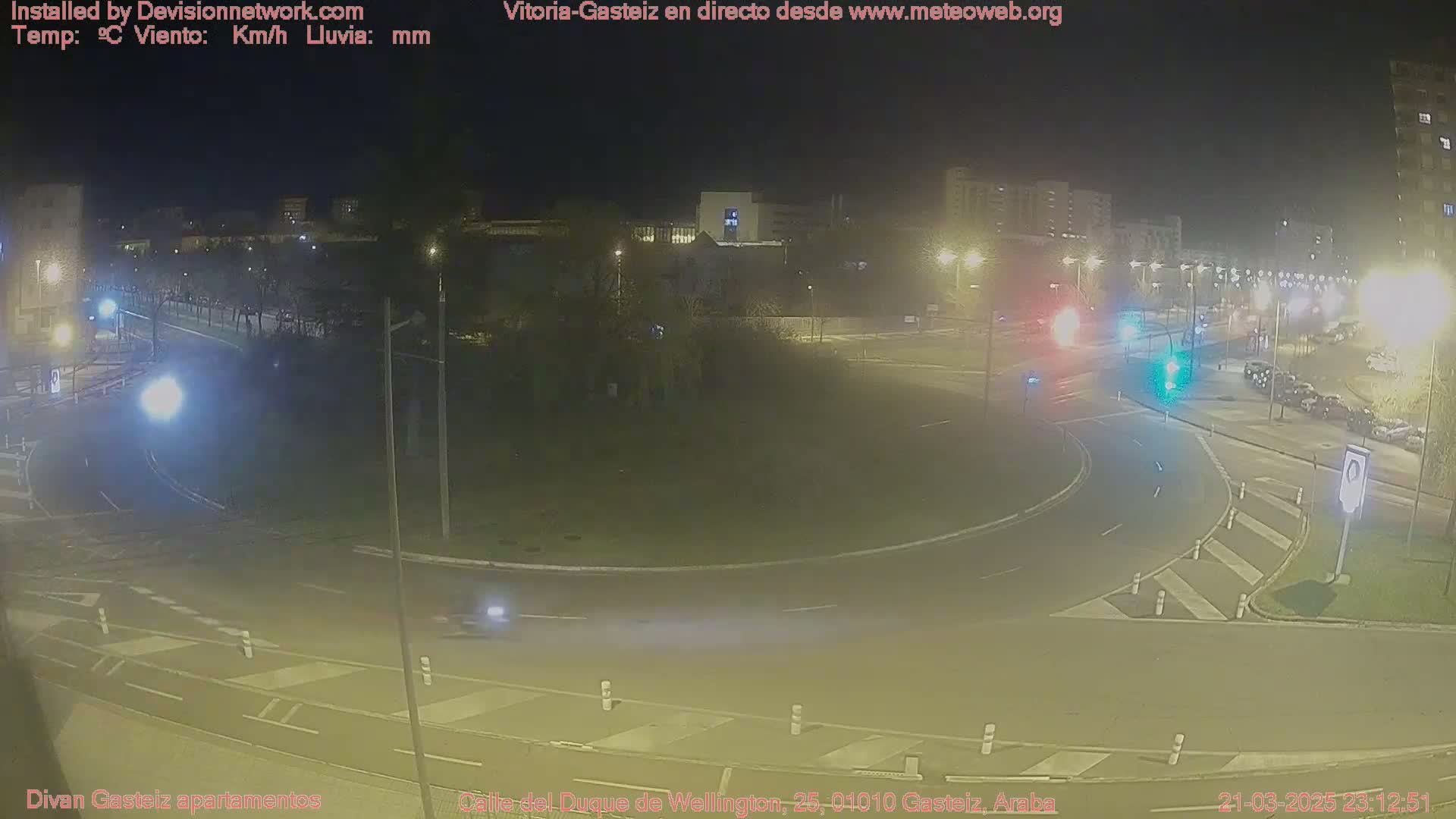 A nighttime view of a roundabout in a city with cars driving on the road and buildings visible in the background under hazy conditions.