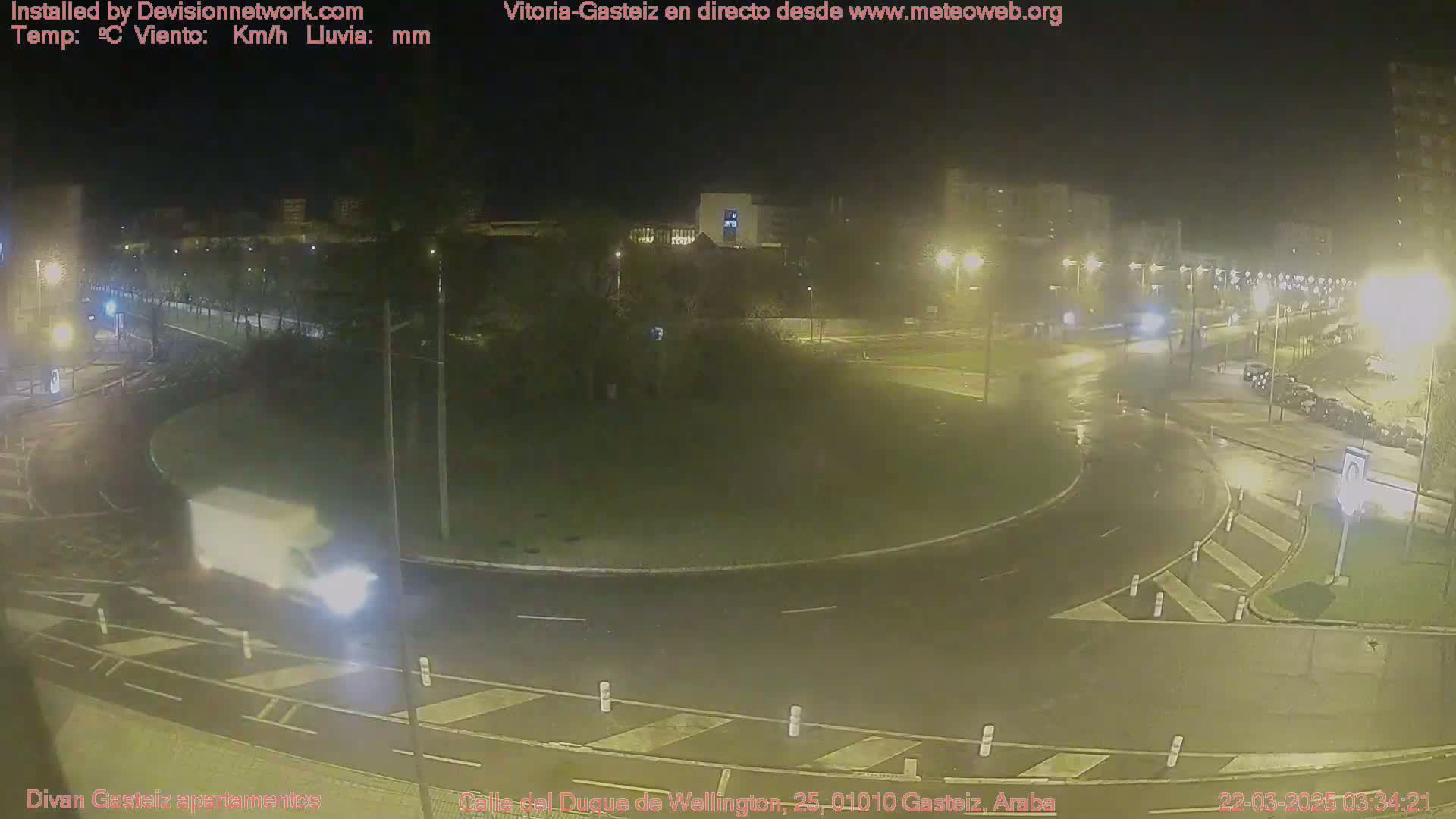 A nighttime, wet roundabout with sparse traffic is illuminated by streetlights.
