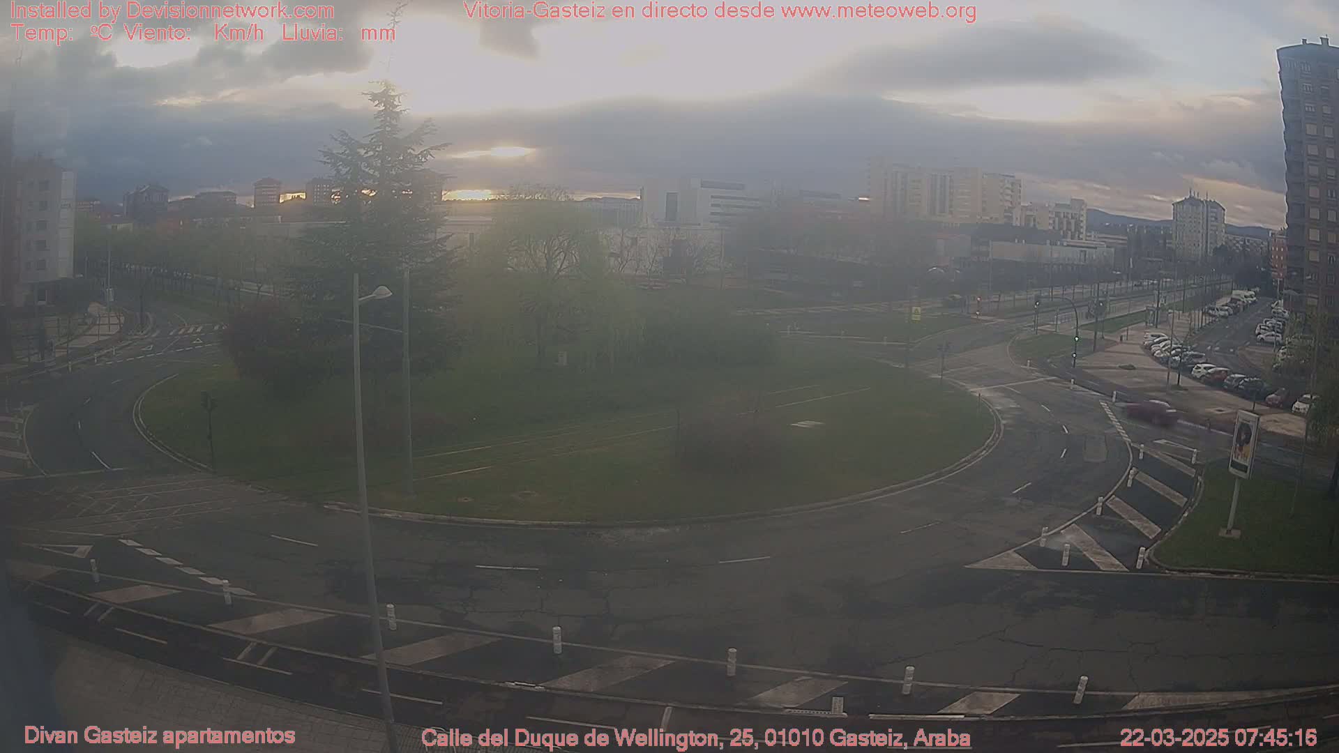 A hazy sunrise illuminates a roundabout and surrounding city streets, with cars parked along the roadside under a mostly cloudy sky.