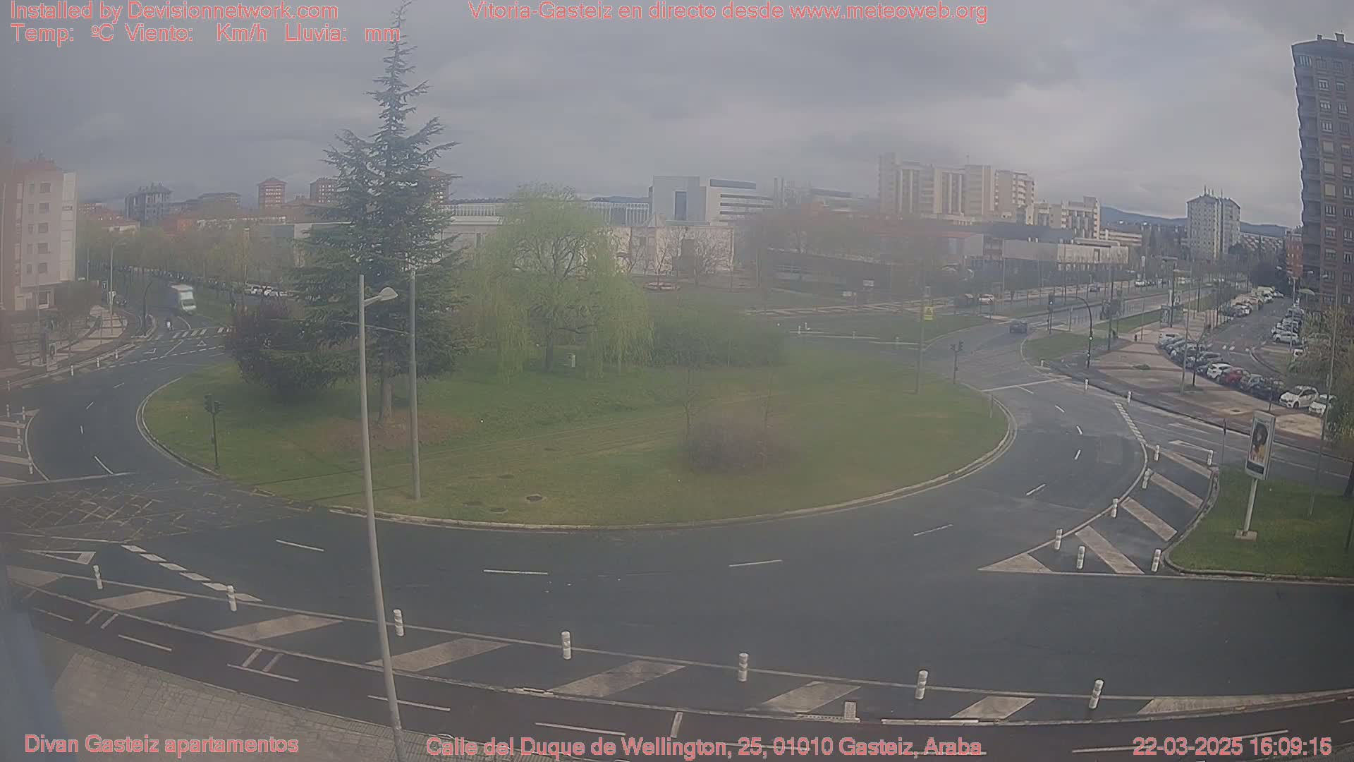A roundabout in a city on an overcast day, with light traffic and several buildings in the background.