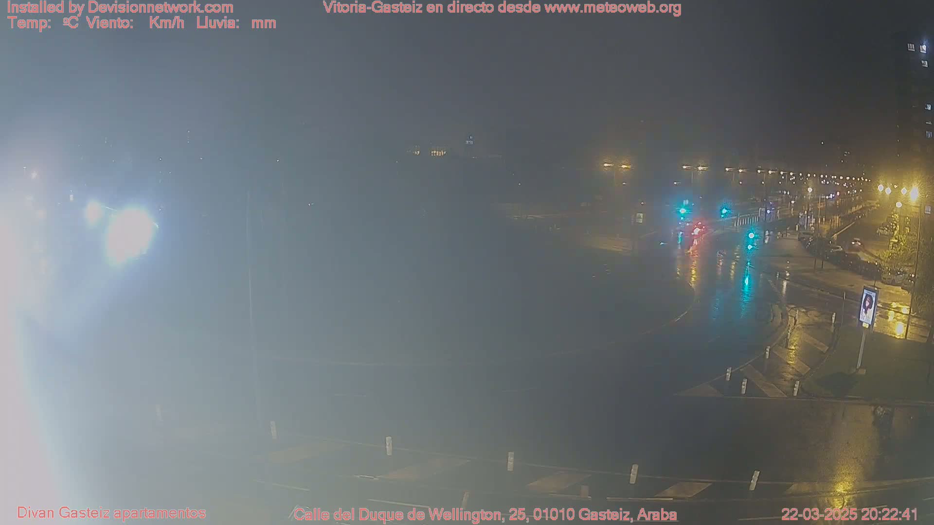 A nighttime, rainy scene shows a wet roadway with sparse traffic and streetlights reflecting on the puddles.