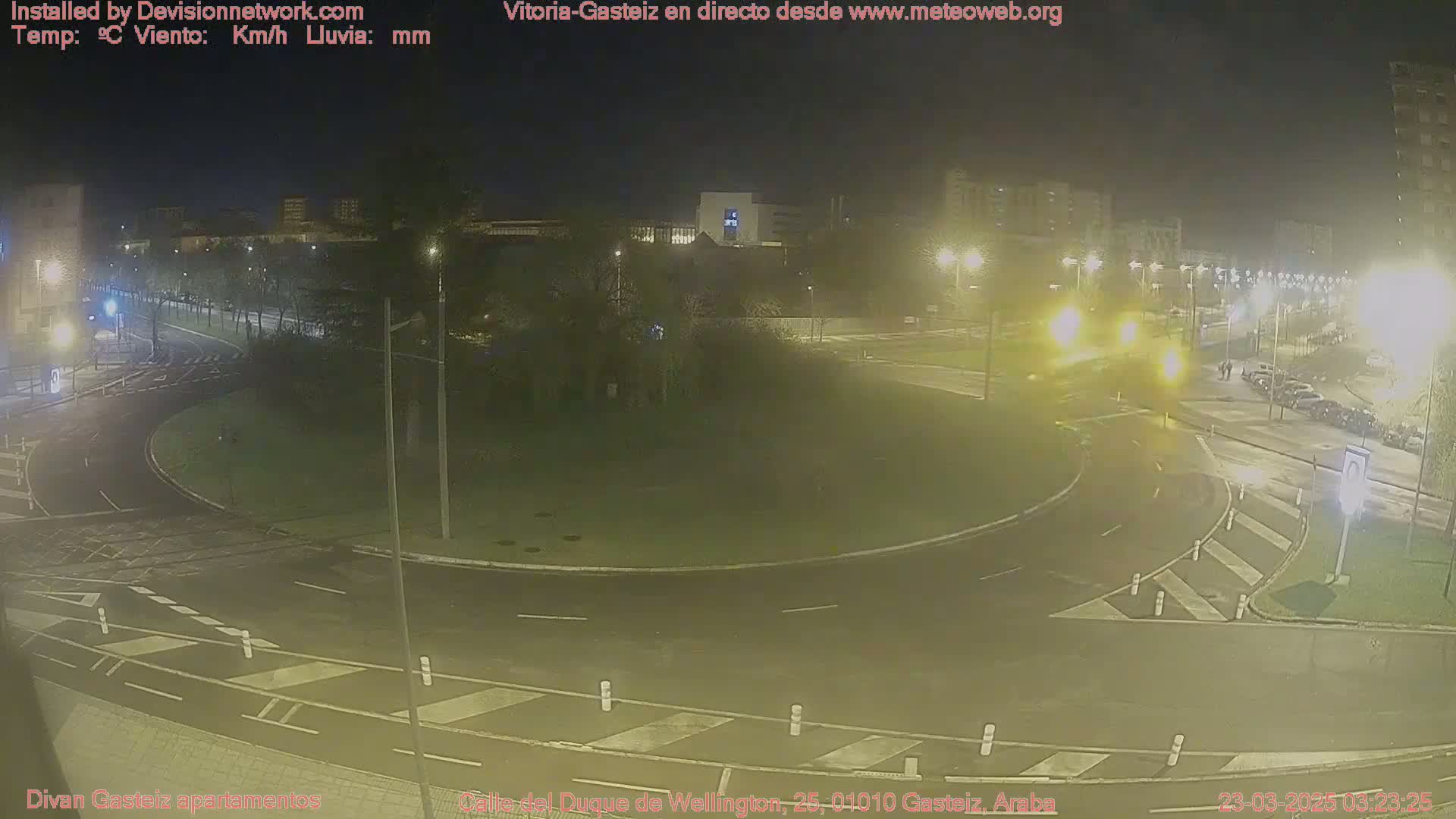 A nighttime view of a roundabout in a city, with light rain on the wet roads and streetlights illuminating the area.