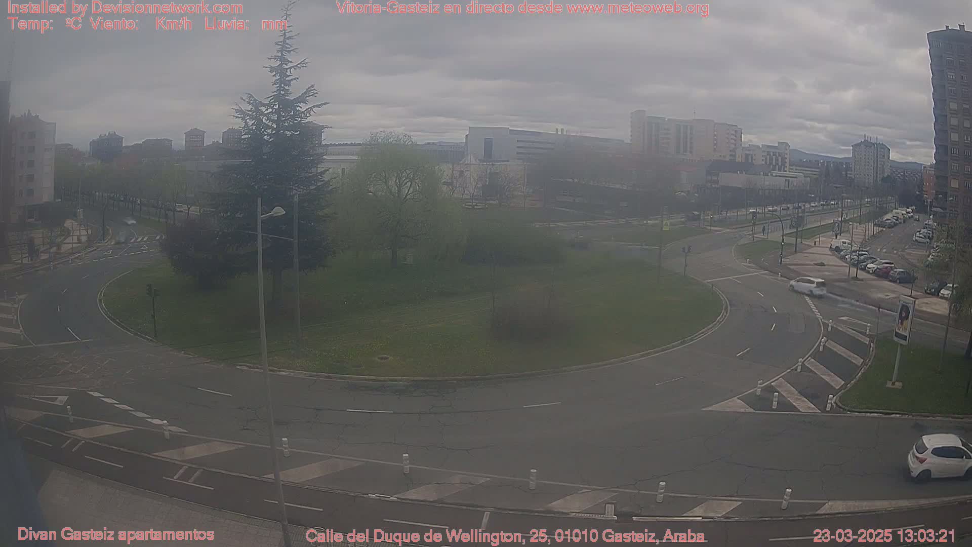 A mostly overcast day shows a roundabout in a city with cars driving around it and buildings in the background.