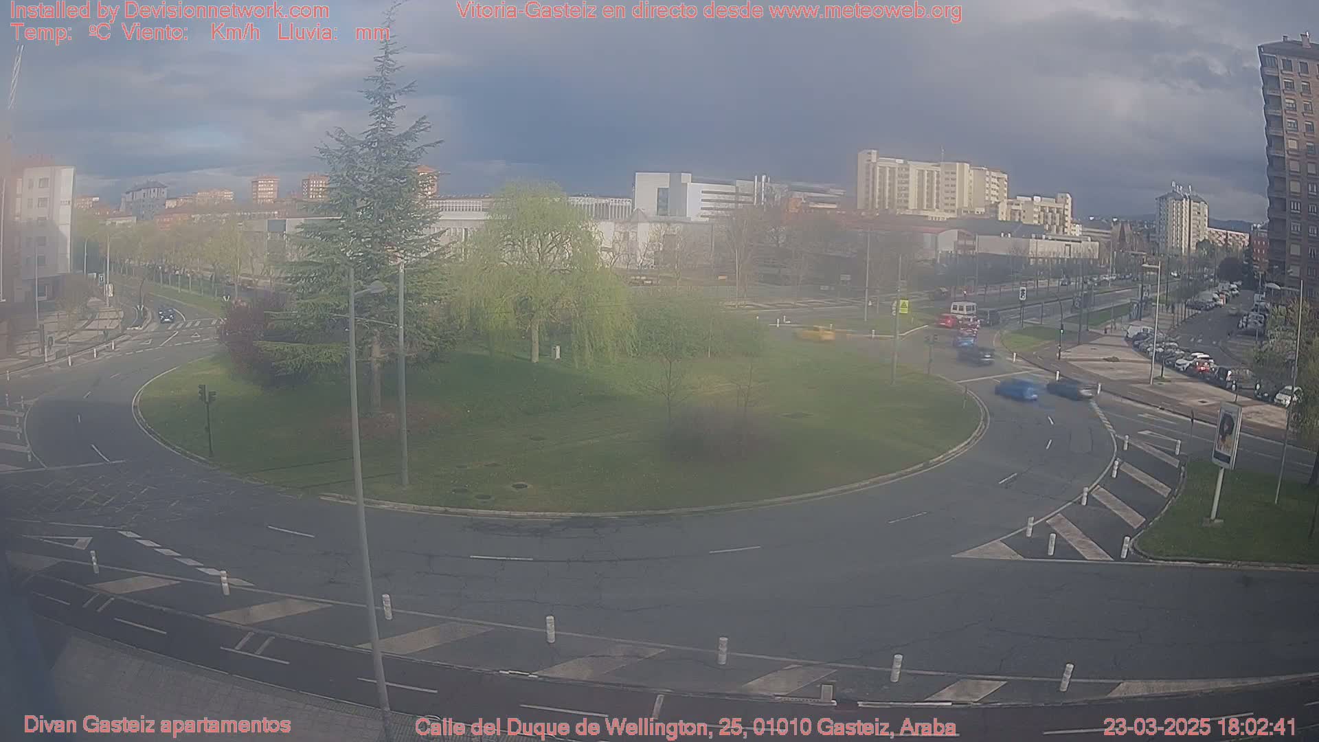 A mostly cloudy day shows a roundabout surrounded by a grassy area, buildings, and several cars moving through the intersection.