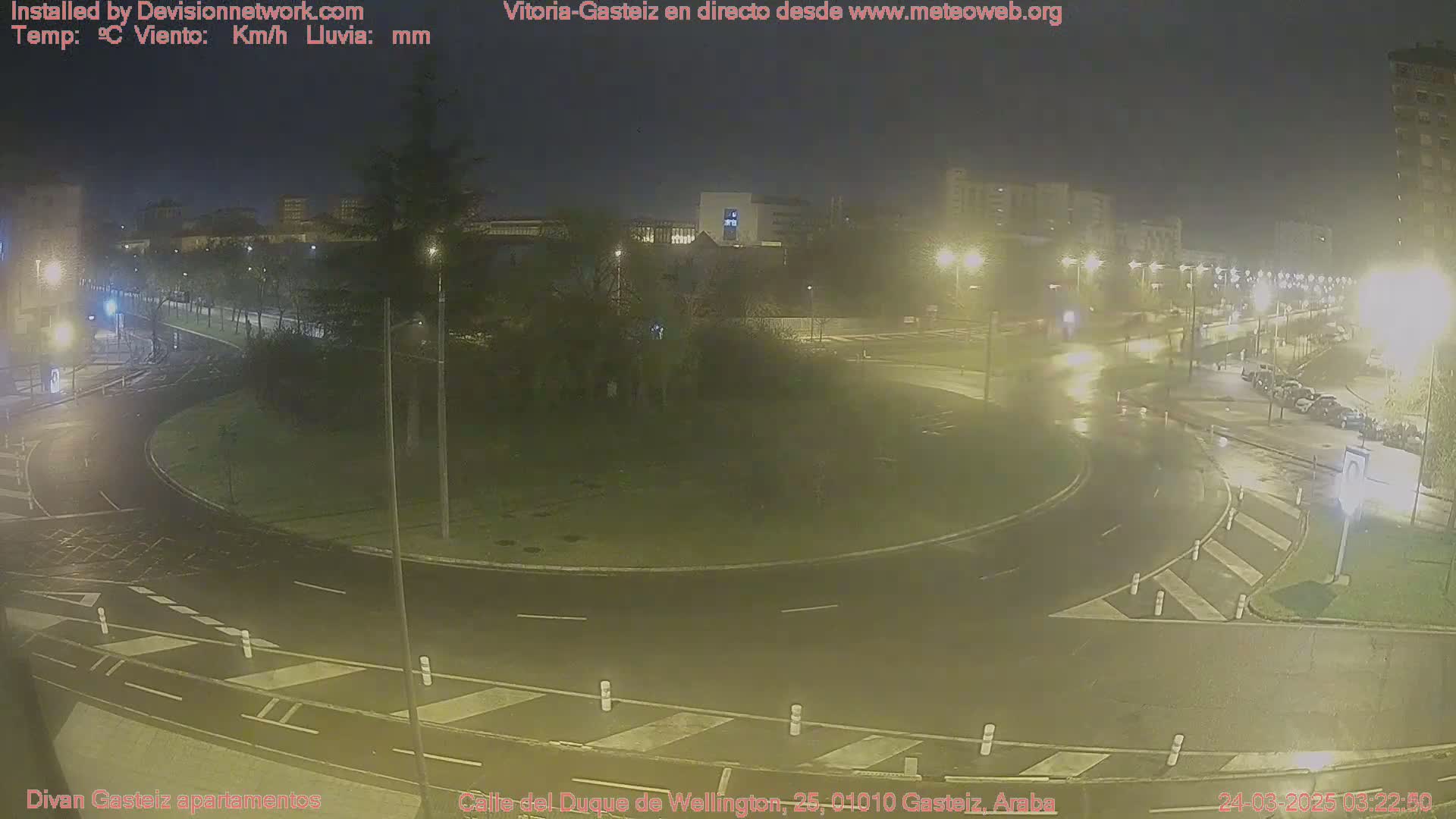A wet, nighttime view of a roundabout in a city with several cars and streetlights.