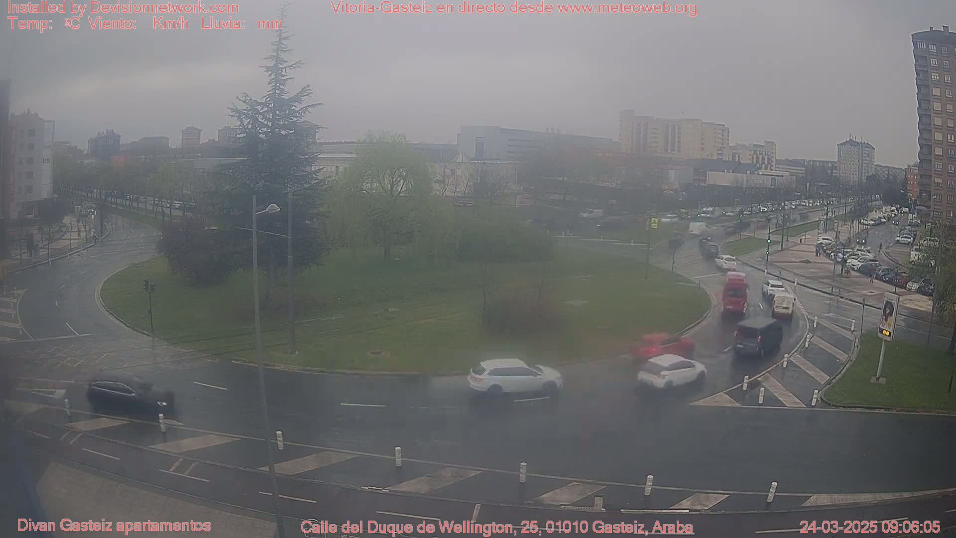 A wet, overcast day shows several cars navigating a roundabout in a city, surrounded by buildings and trees.