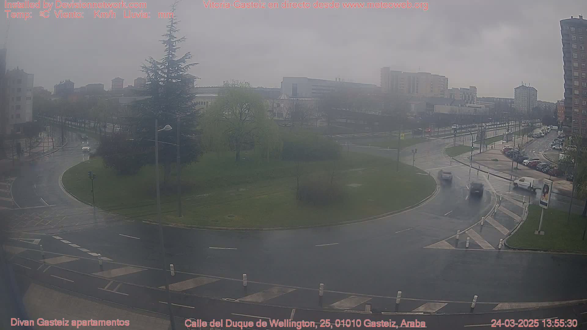 A rainy scene shows a roundabout in a city with cars driving on wet roads, surrounded by buildings and trees under a gray sky.