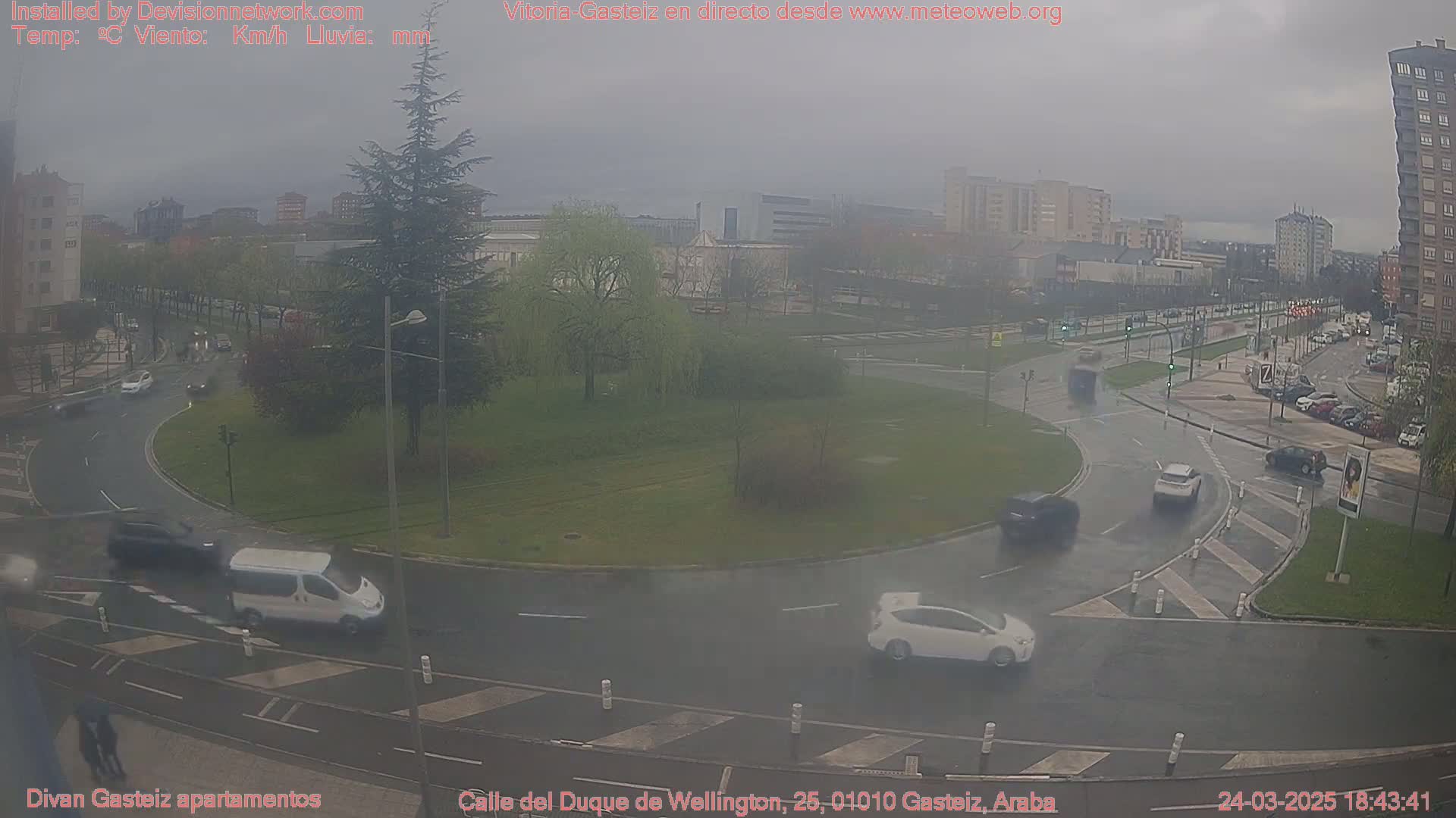 A wet roundabout in a city with several cars driving on it, surrounded by buildings and trees under an overcast sky.