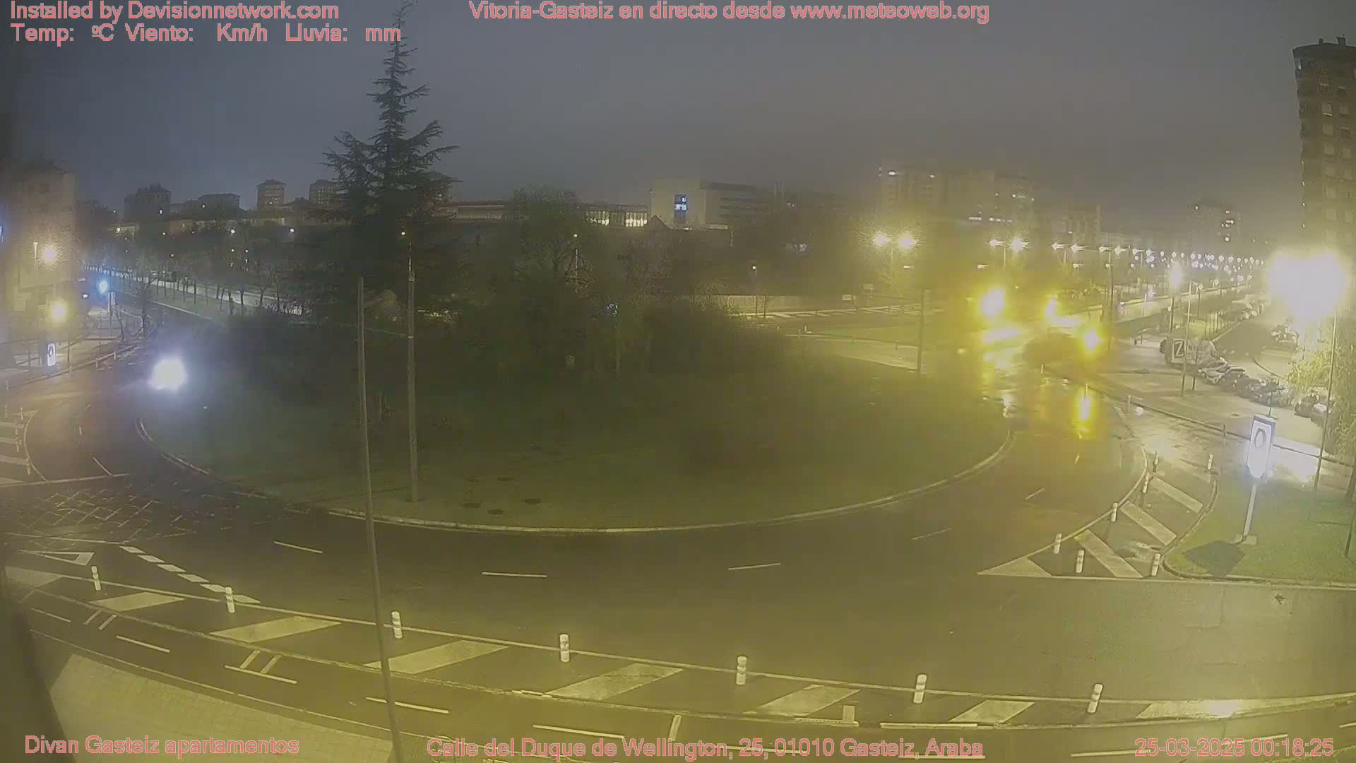 A wet, nighttime scene shows a roundabout with cars and streetlights illuminated in the rain.