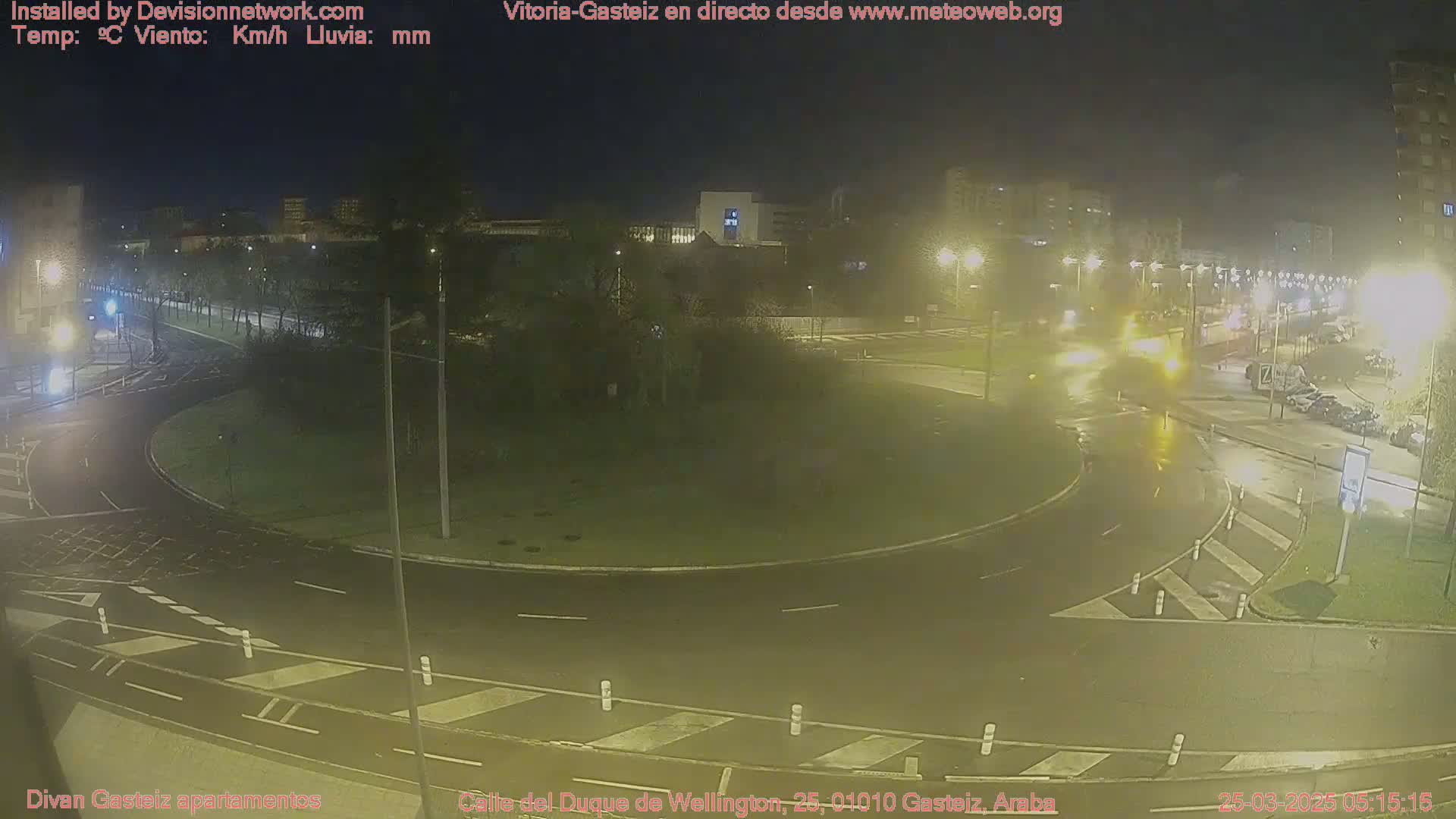 A nighttime view of a wet roundabout and surrounding roads in a city, with buildings and streetlights visible in the background.
